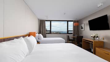 Quarto de hotel com duas camas, uma escrivaninha, uma cadeira, uma televisão e janela com vista para a cidade.