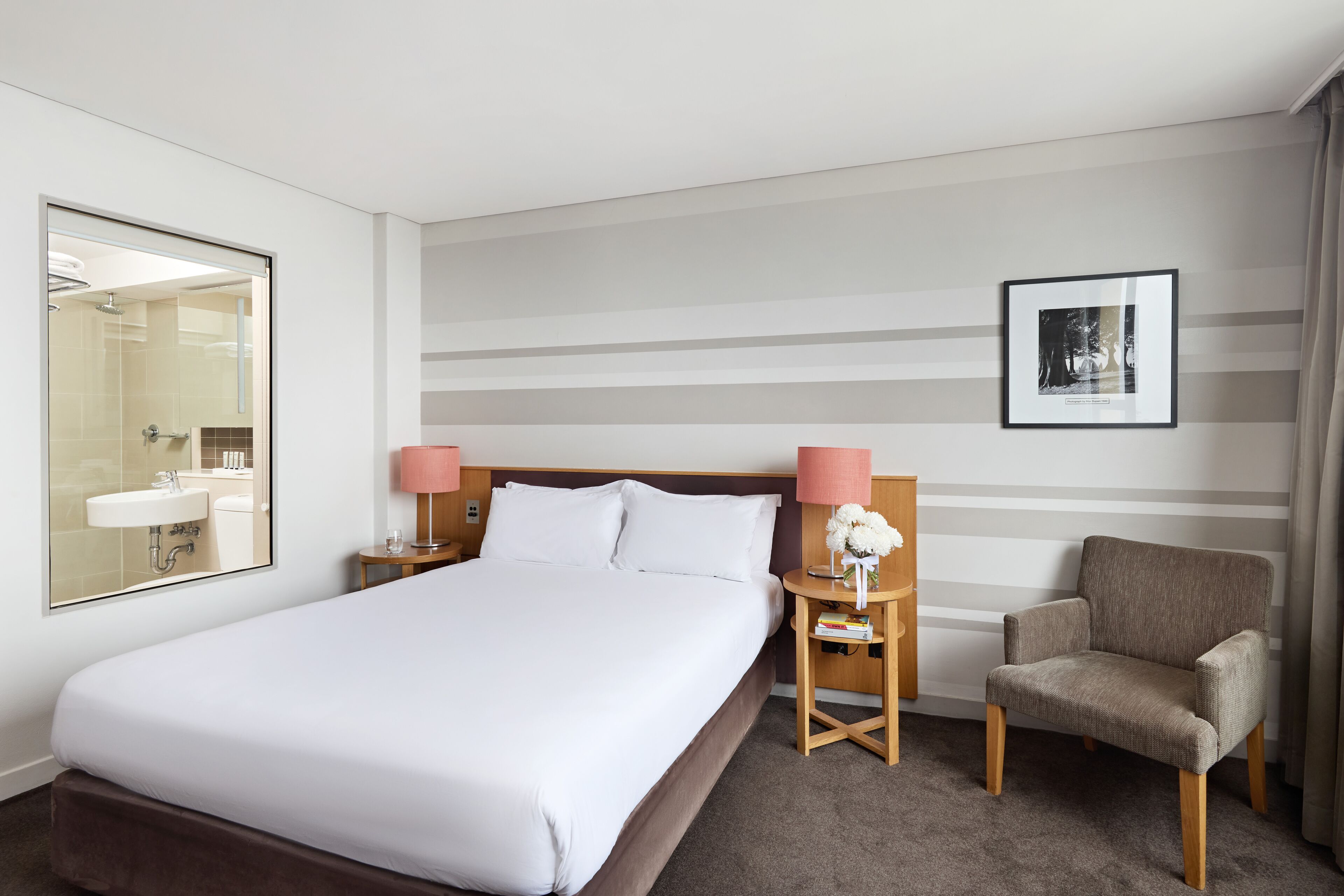 A hotel room with a large bed, a bedside table with a lamp, a chair, a framed picture on the wall, and a mirror.
