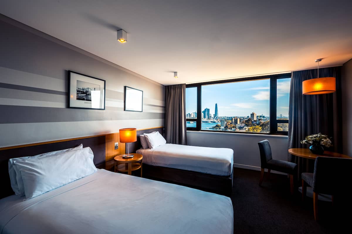 A hotel room with two beds, a desk, a chair, a lamp, a framed picture, and a view of the cityscape.