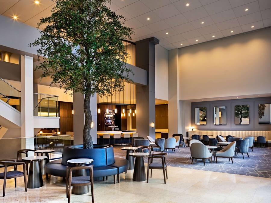 A spacious lobby with a central tree, multiple seating areas, a bar, and a staircase.