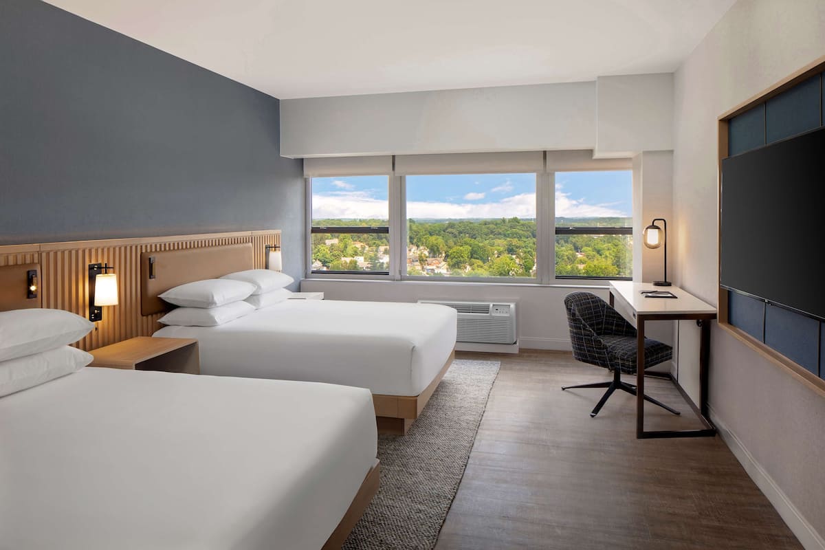 Habitación de hotel con dos camas, un escritorio, una silla y un ventanal con vistas a la ciudad.
