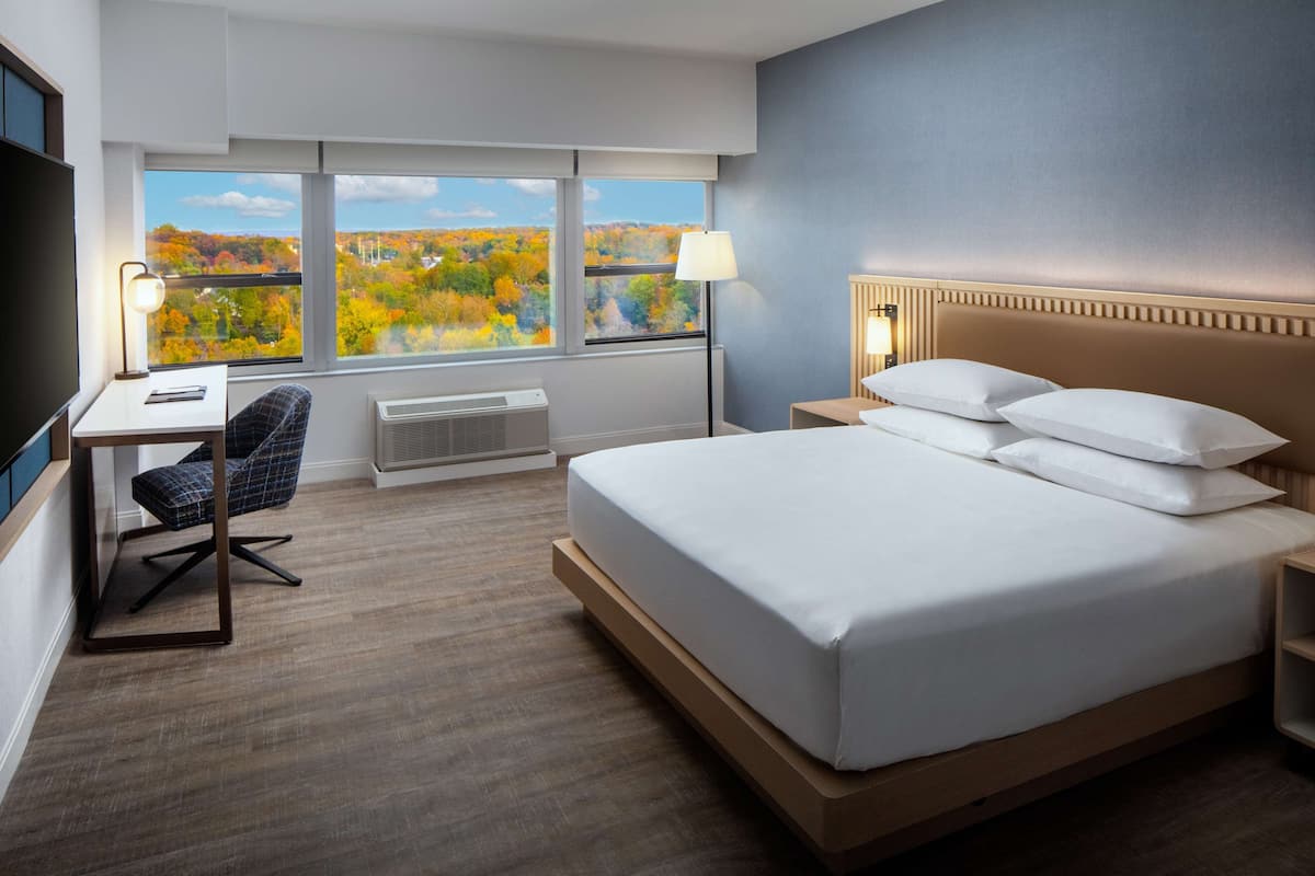 Habitación de hotel con una cama grande, un escritorio, una silla y vistas a la arboleda otoñal a través de las ventanas.
