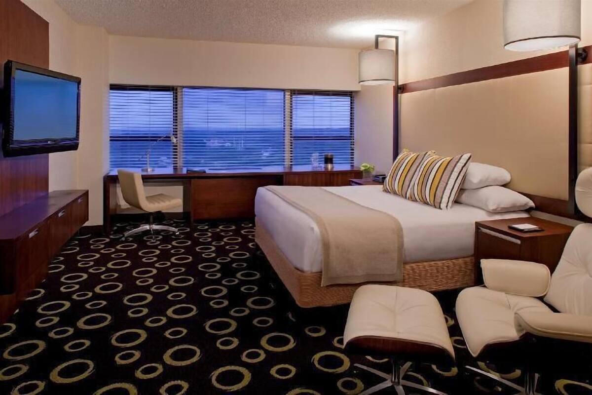 A hotel room with a bed, desk, chair, TV, and large windows.