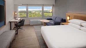 Habitación de hotel con cama, escritorio, silla, mesita pequeña y sofá. Amplias ventanas ofrecen vistas a zonas verdes y edificios.