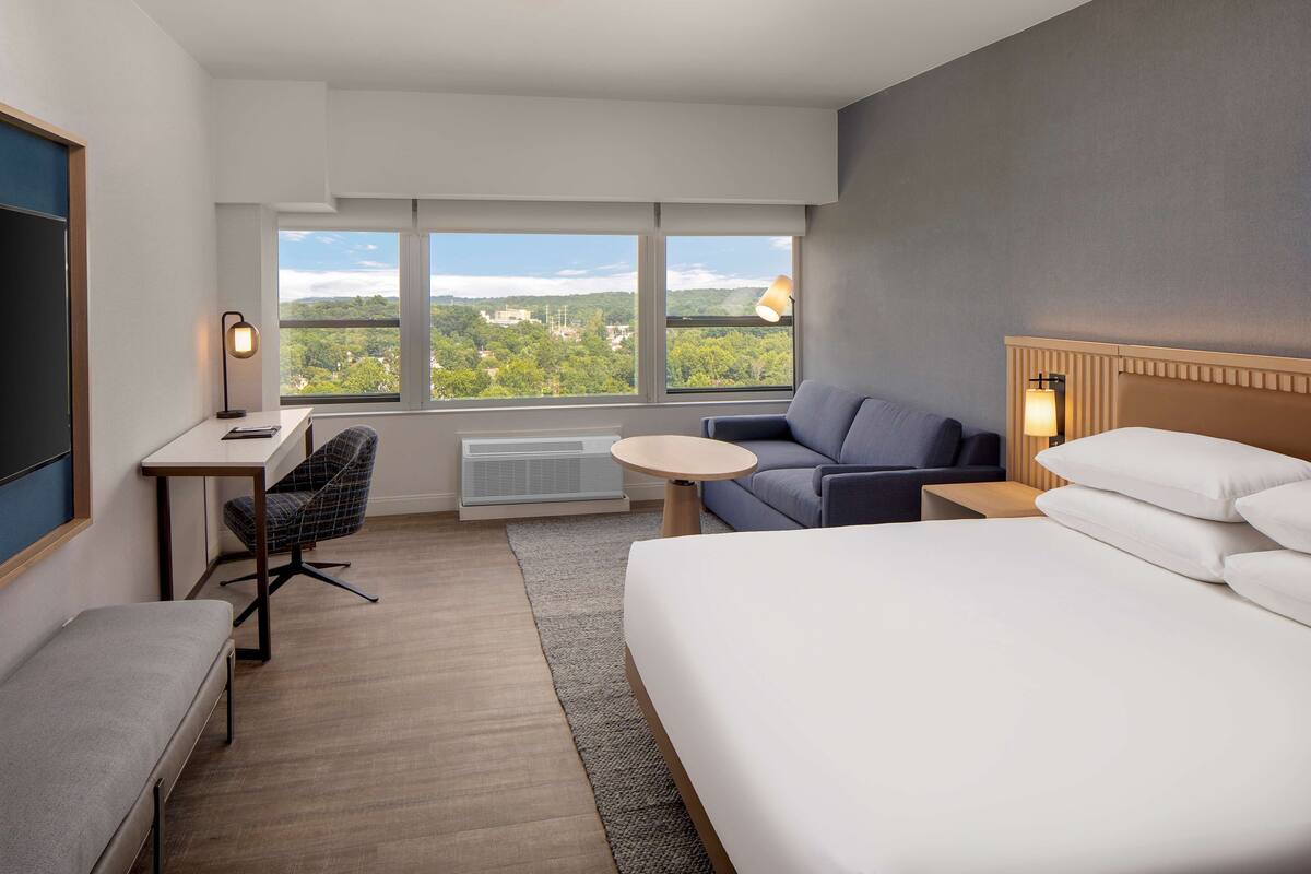 Habitación de hotel con cama, escritorio, silla, mesita pequeña y sofá. Amplias ventanas ofrecen vistas a zonas verdes y edificios.