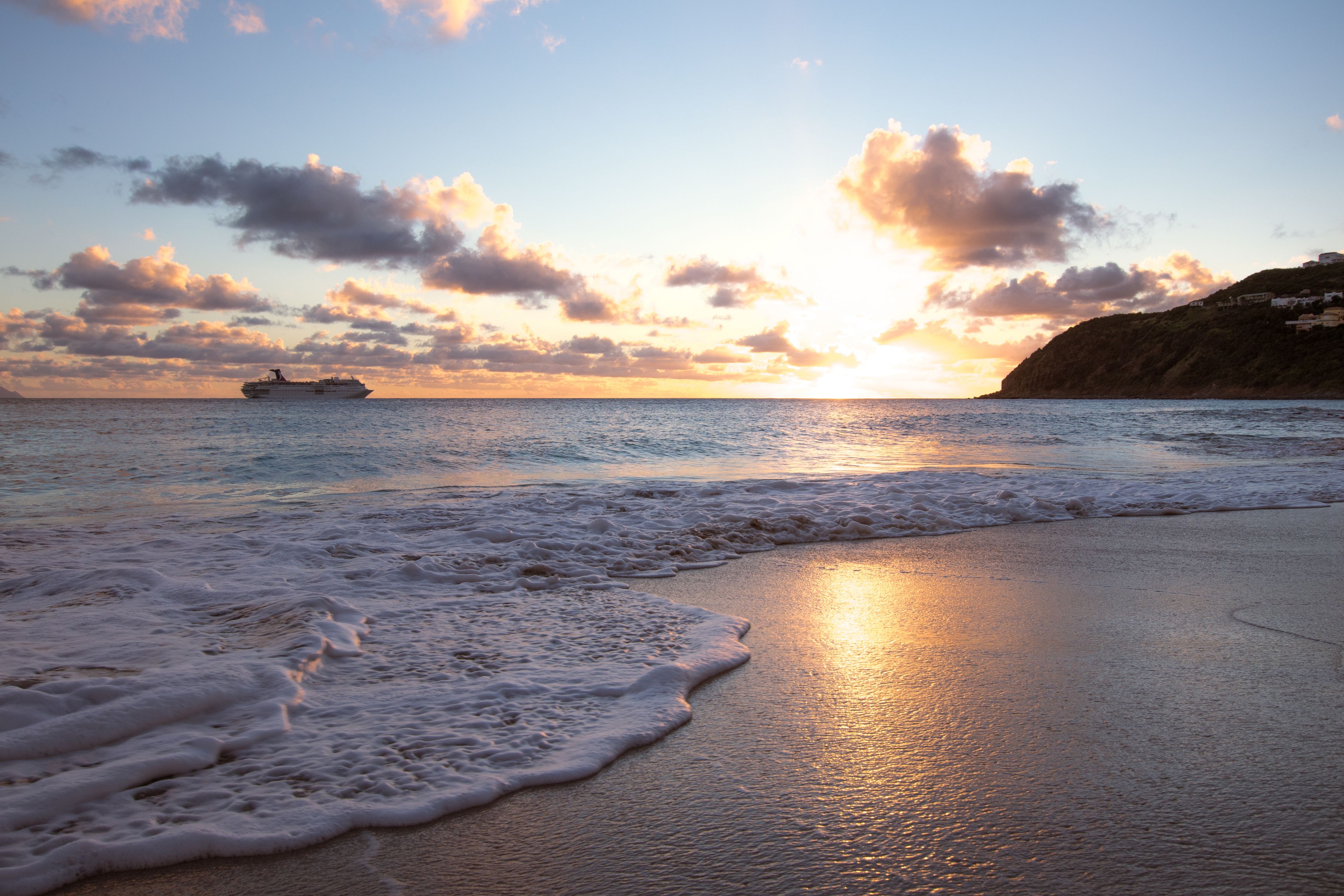 A beach with waves at sunset, a ship in the distance, and a cliff with buildings.
