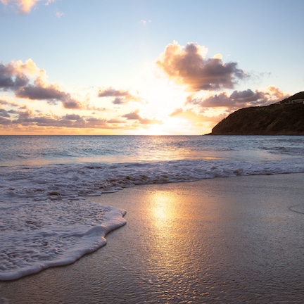 A beach with waves at sunset, a ship in the distance, and a cliff with buildings.