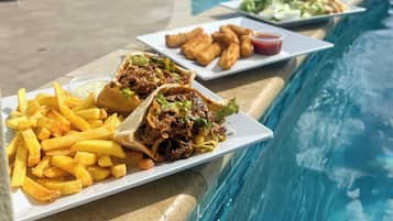 A poolside dining area with a view of the sea, featuring plates of food including fries, tacos, and fried items, and a pool with clear water.