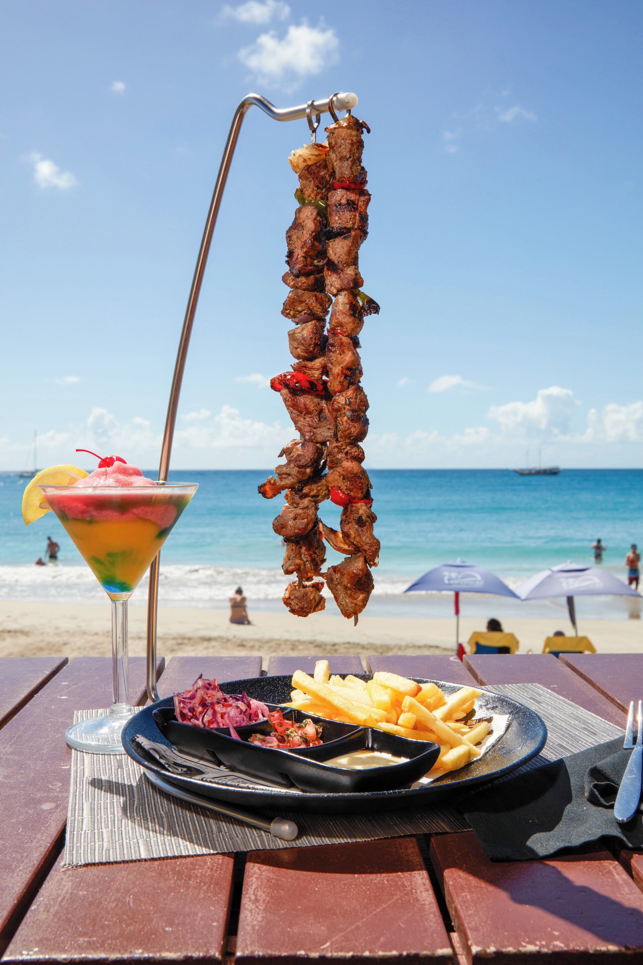 Un montaje para comer frente al mar, con un pincho de carne, una porción de papas fritas y una bebida de colores en una mesa.