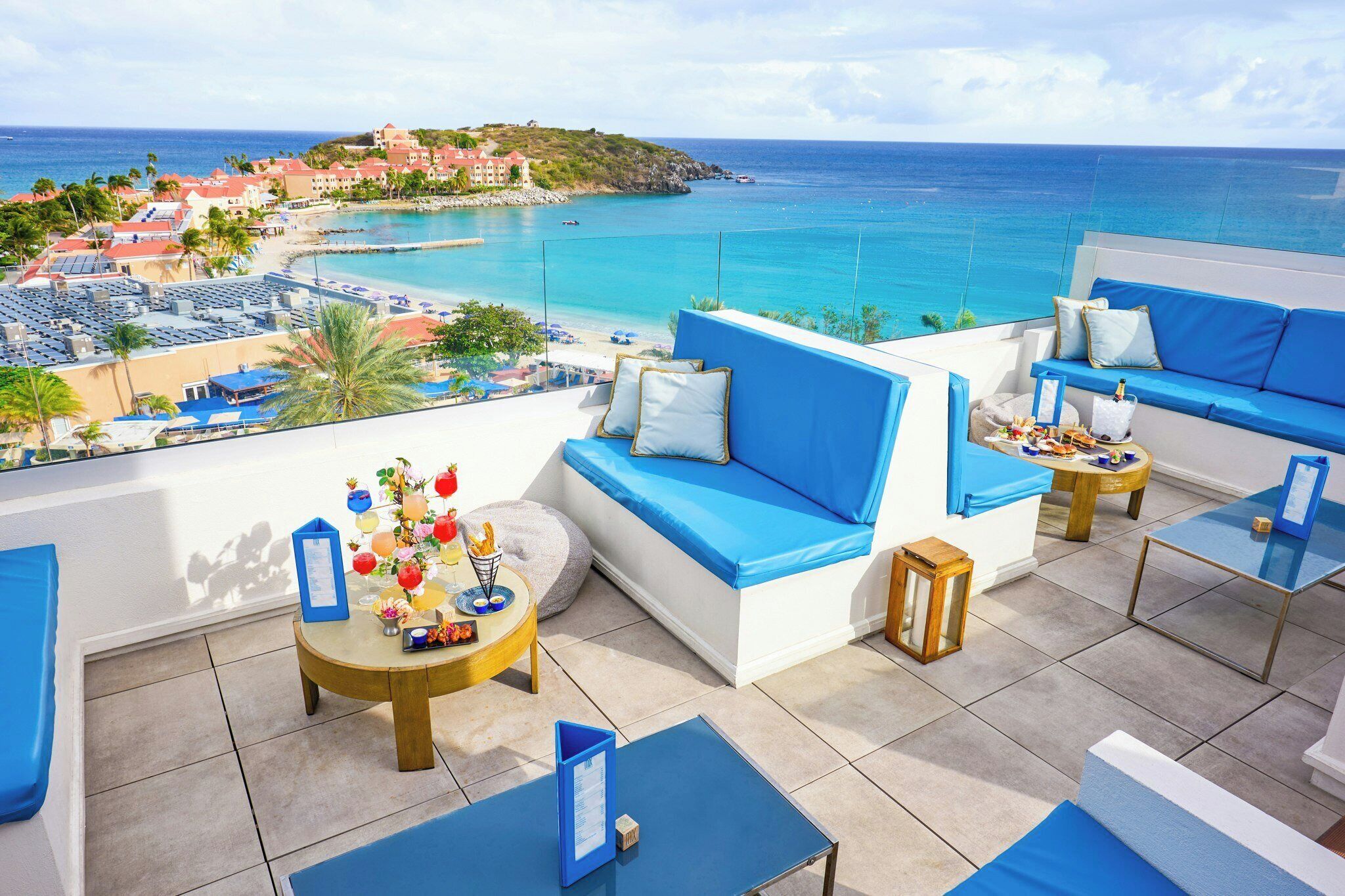 A rooftop terrace with blue cushioned seating, a round table with a drink menu, and a view of the ocean and resort buildings.