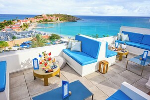 A rooftop terrace with blue cushioned seating, a round table with a drink menu, and a view of the ocean and resort buildings.