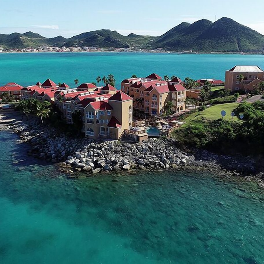 An aerial view of a coastal resort with buildings featuring red roofs, situated near clear turquoise waters and a cruise ship in the background.