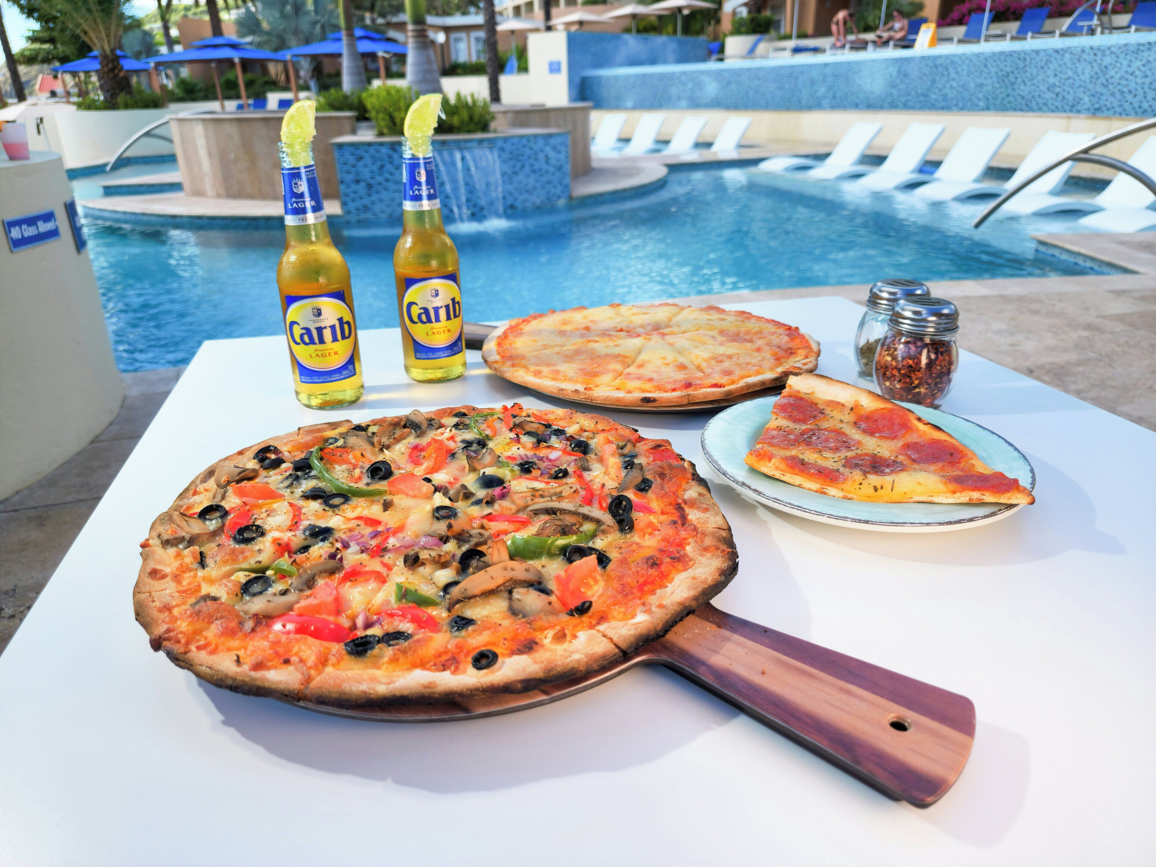Une table avec deux pizzas, une part de pizza sur une assiette et deux bouteilles de bière près d’une piscine.