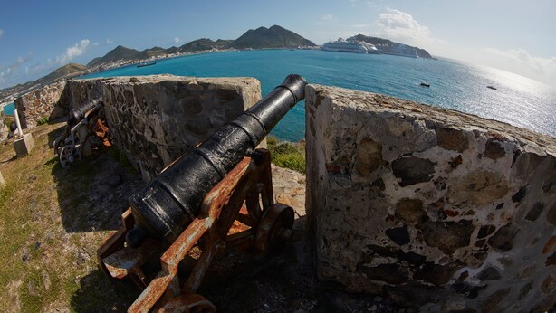 A cannon positioned on a stone platform overlooking a coastal area with clear turquoise waters and distant mountains.