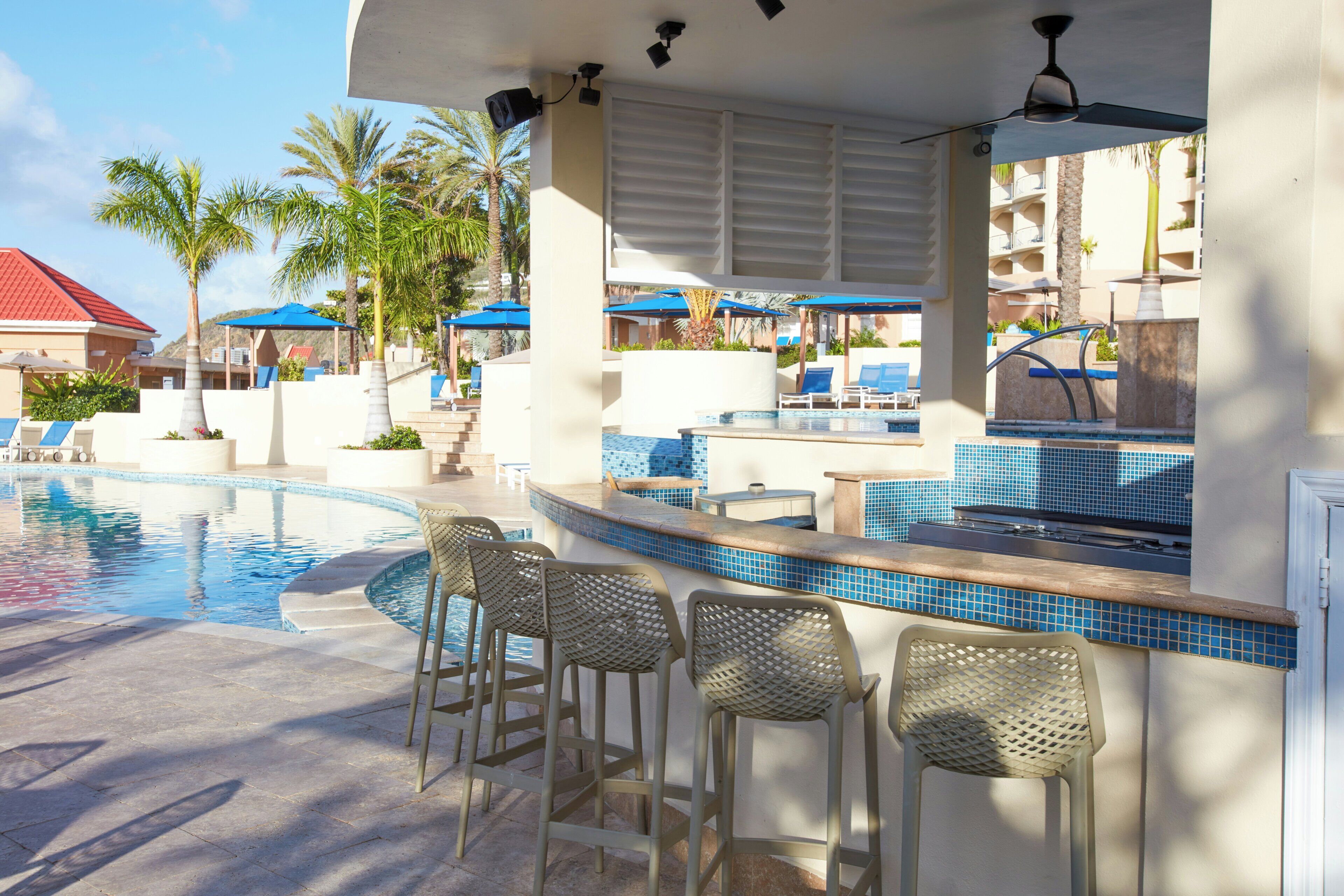 Zona de bar junto a la piscina, con piscina, taburetes y sombrillas.