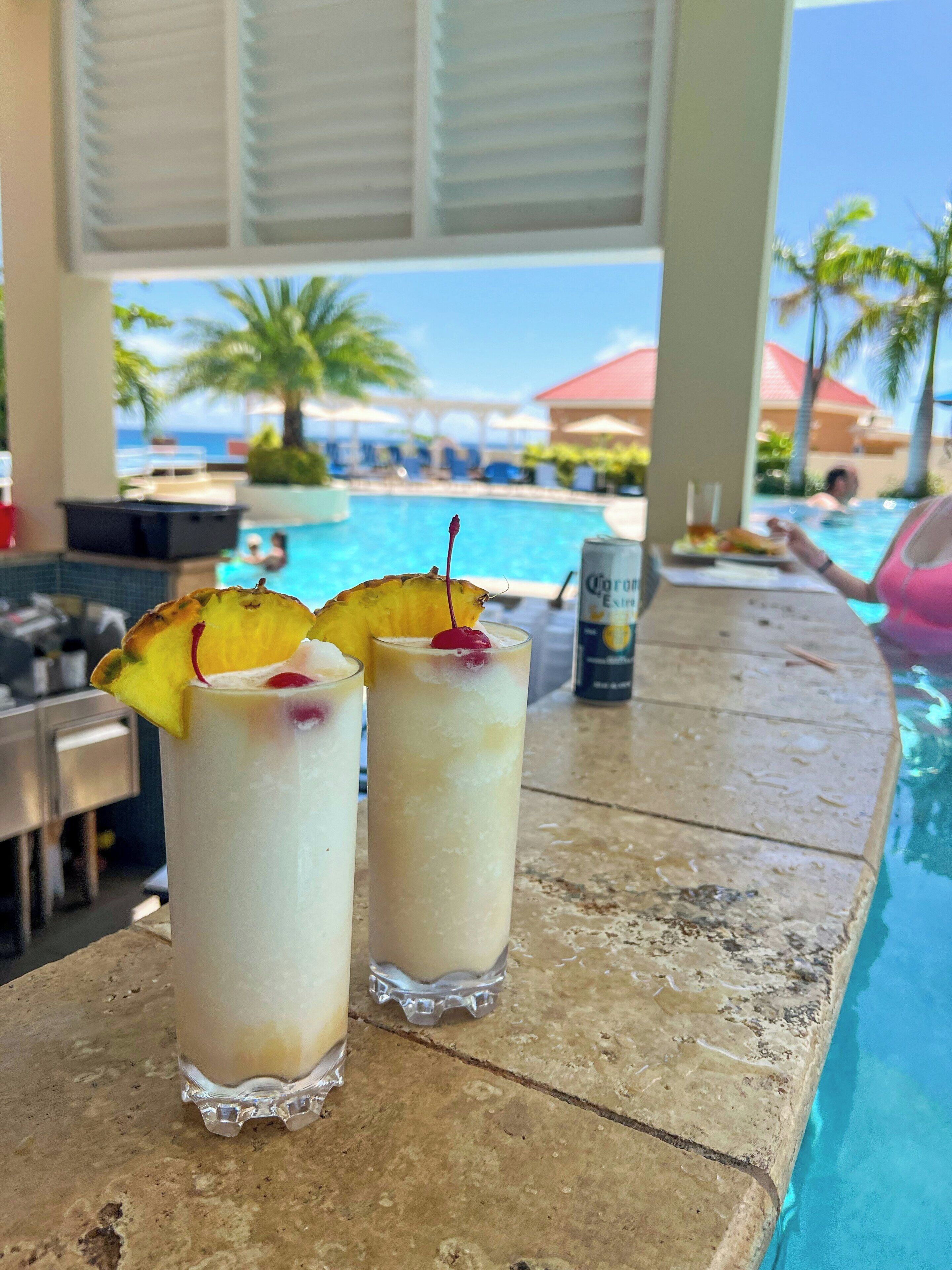 Two tall glasses of a creamy beverage with fruit garnishes on a poolside bar, with a pool and palm trees in the background.
