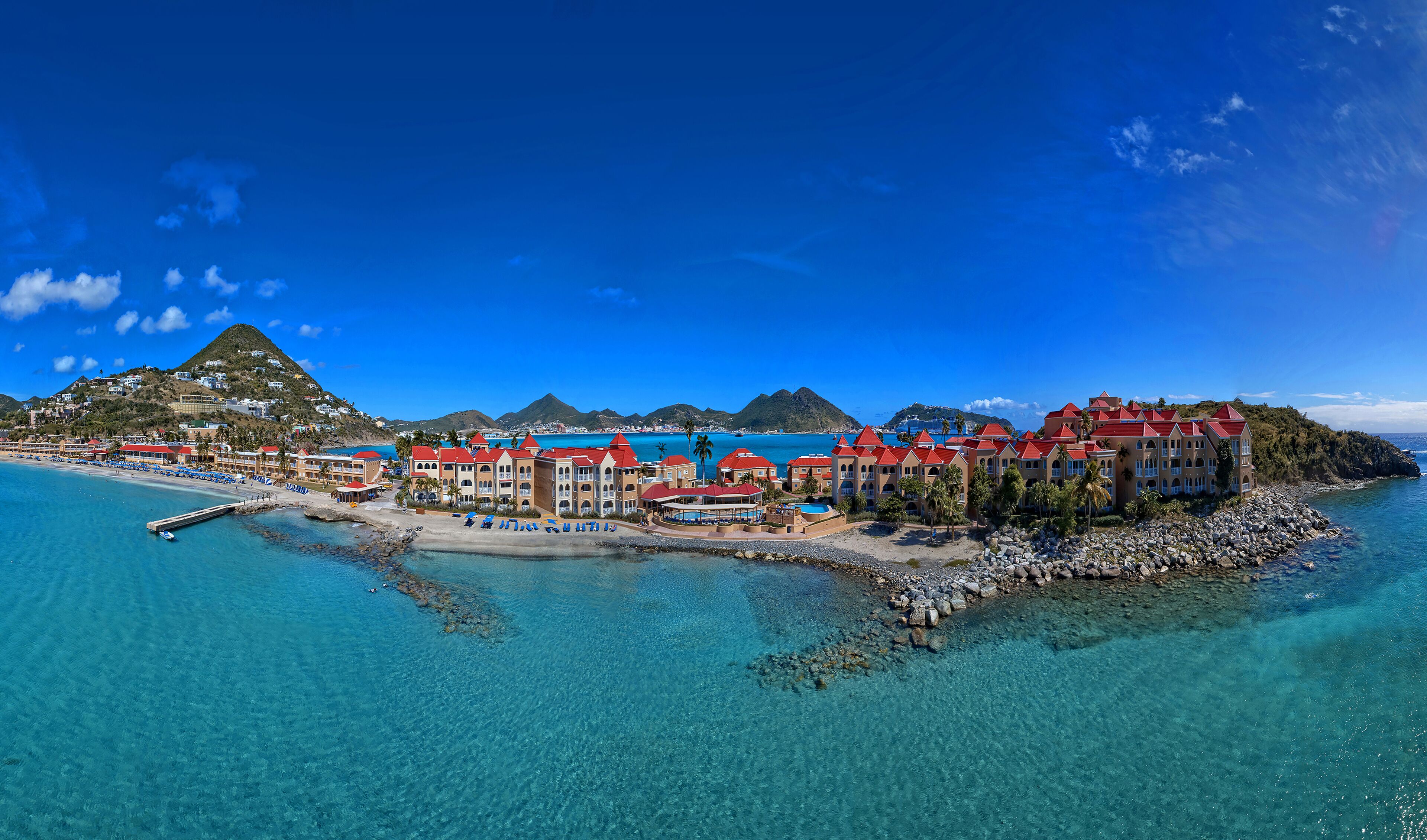 A coastal resort with multiple buildings, clear blue waters, and a mountainous backdrop.