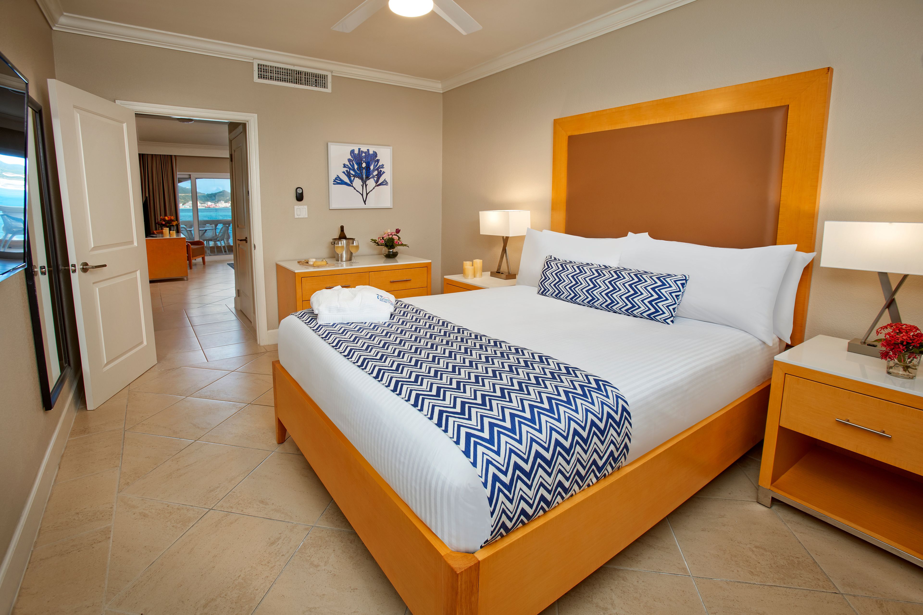 Une chambre d’hôtel avec un grand lit, une tête de lit en bois, deux tables de chevet avec des lampes, une petite table et une vue sur la plage par une porte ouverte.