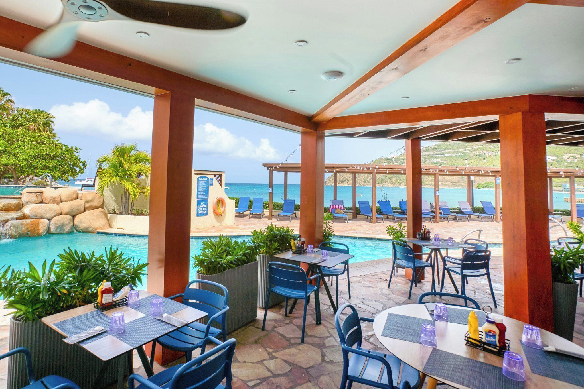 An outdoor pool area with lounge chairs and tables, a view of the sea, and a rocky landscape.