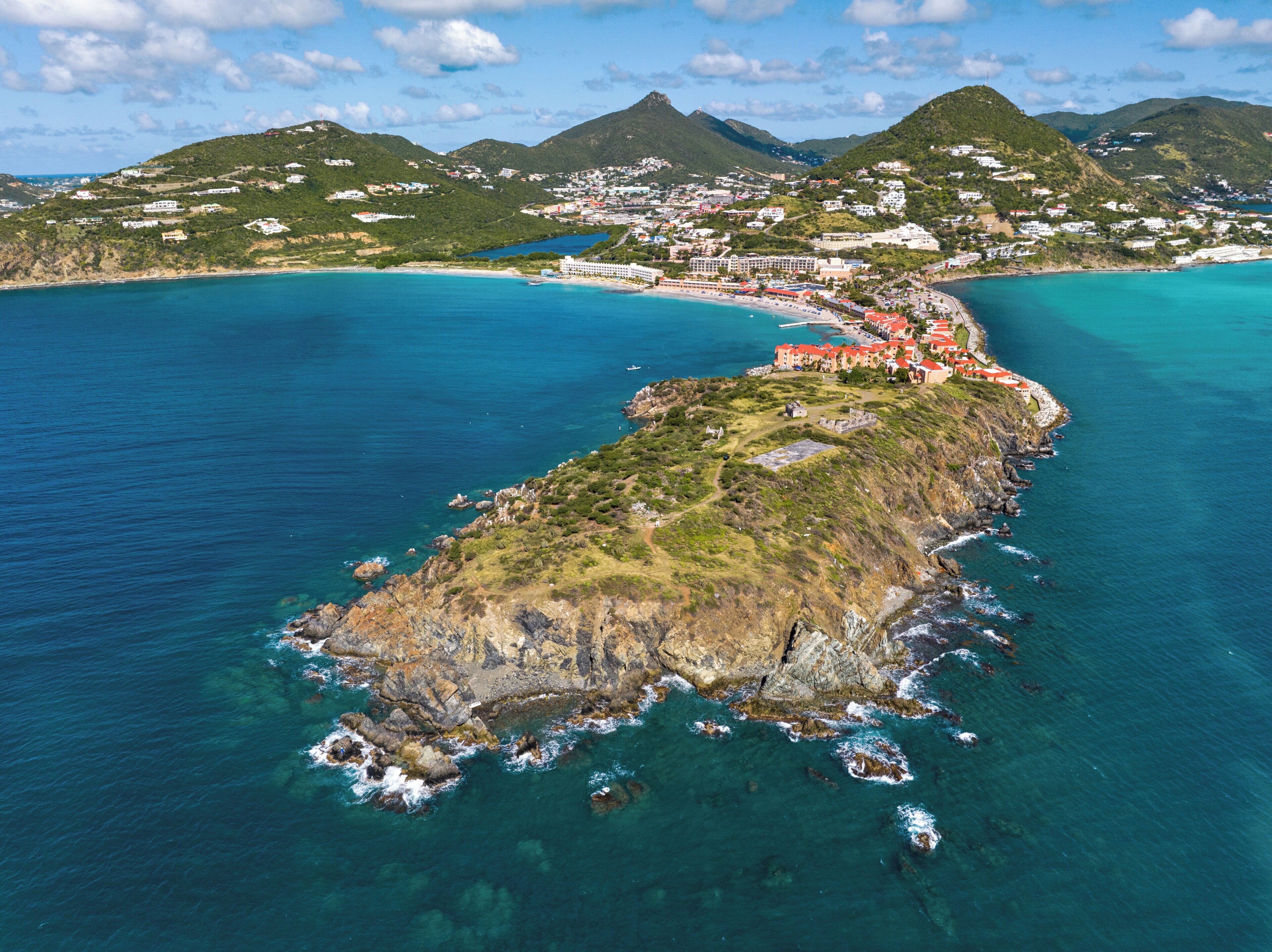 An aerial view of a coastal area with a prominent peninsula, clear blue waters, and a town with buildings on the peninsula.