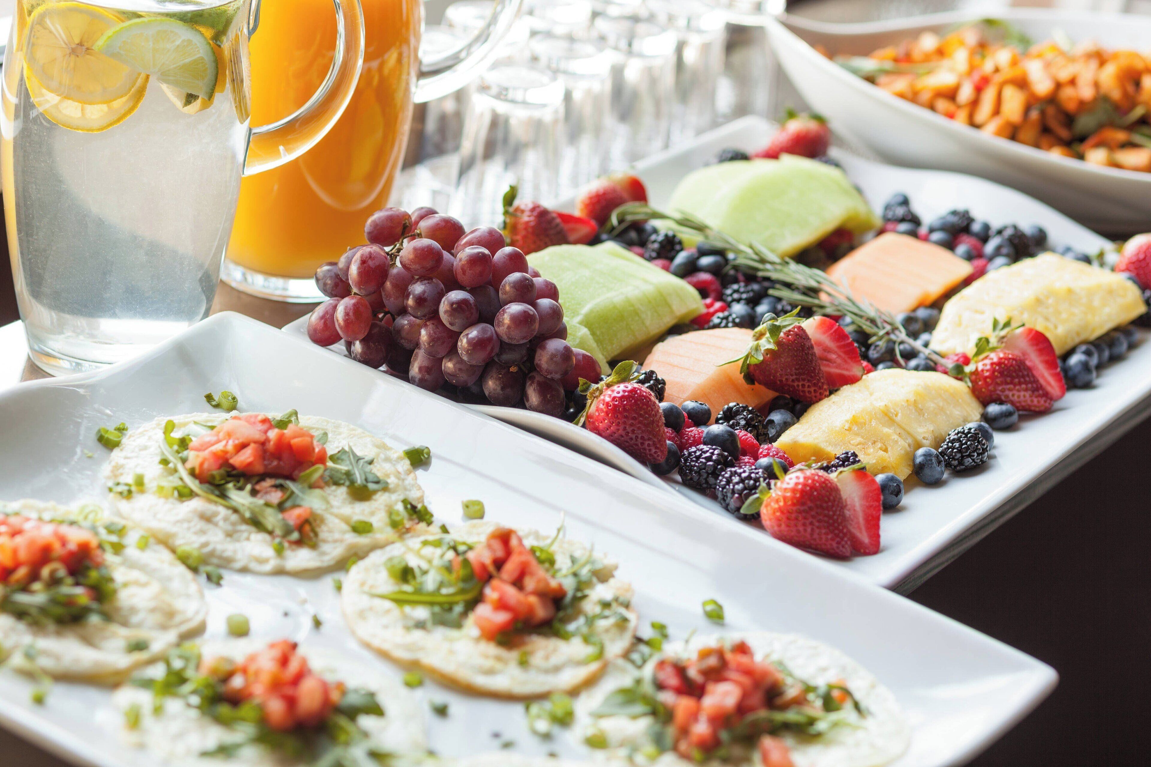 A breakfast buffet with various fruits, cheeses, and a pitcher of orange juice.