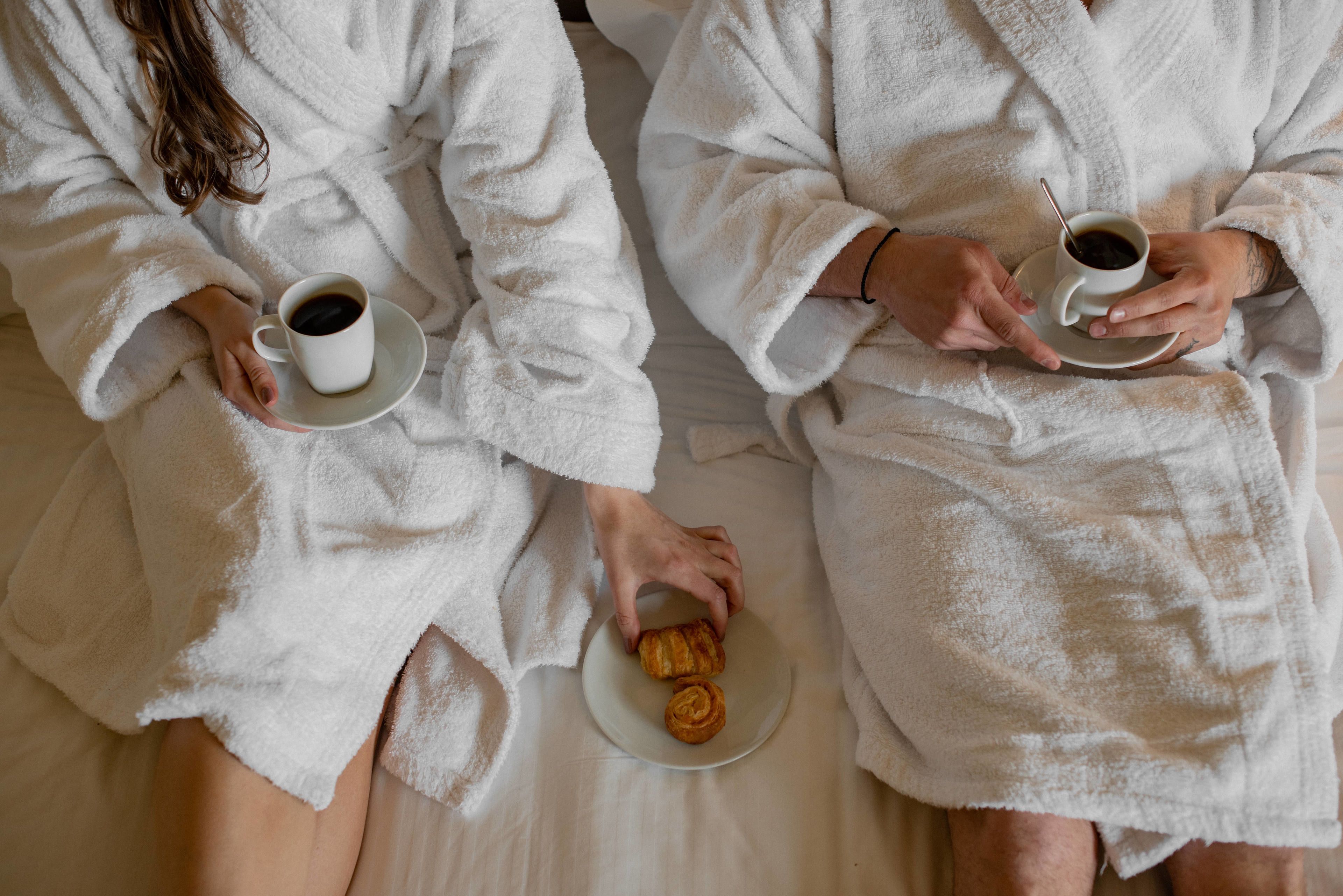 Zwei Personen in weißen Bademänteln sitzen auf einem Bett, jede mit einer Tasse Kaffee und einem Tablett mit Croissants.