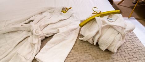 A neatly made bed with white linens, a beige quilted bedspread, and two white bathrobes on a wooden floor.