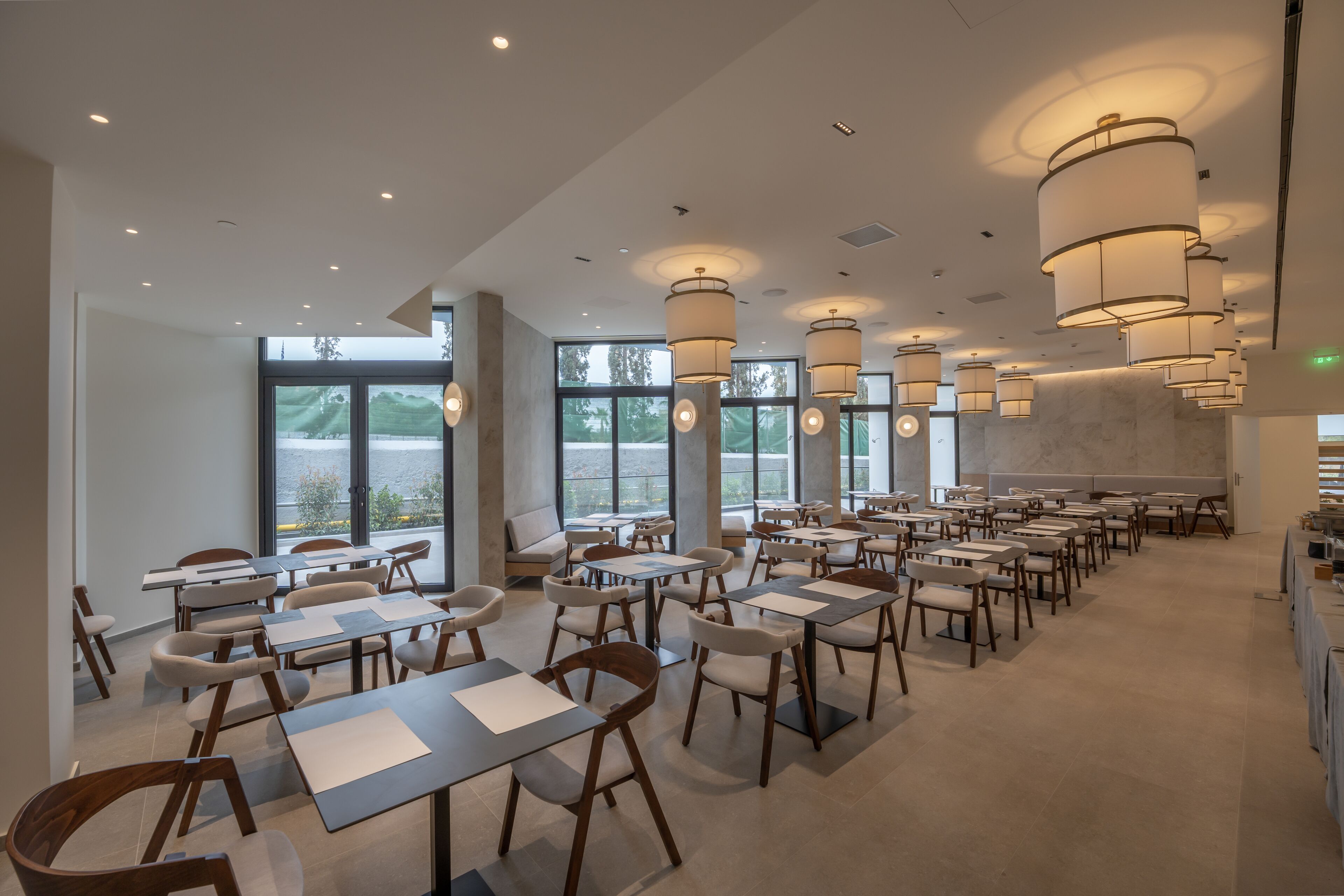 A spacious restaurant with high ceilings, modern pendant lighting, and large windows offering a view of the outdoors.