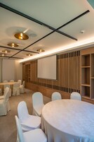Un salón de conferencias con mesas redondas, sillas blancas, una pantalla de proyector y un panel de pared de madera.