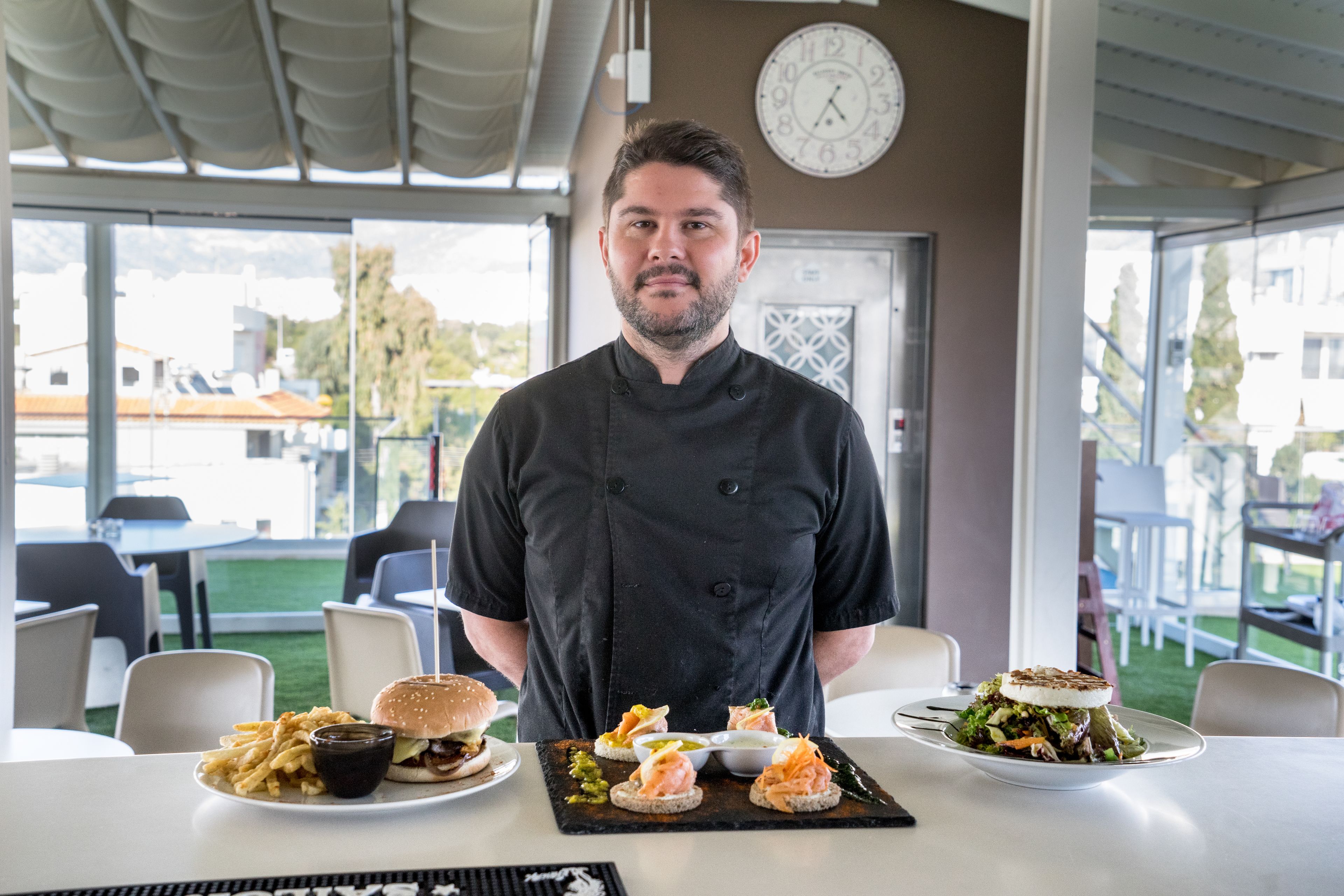 A chef standing behind a table with various dishes, including a burger, fries, and a salad.