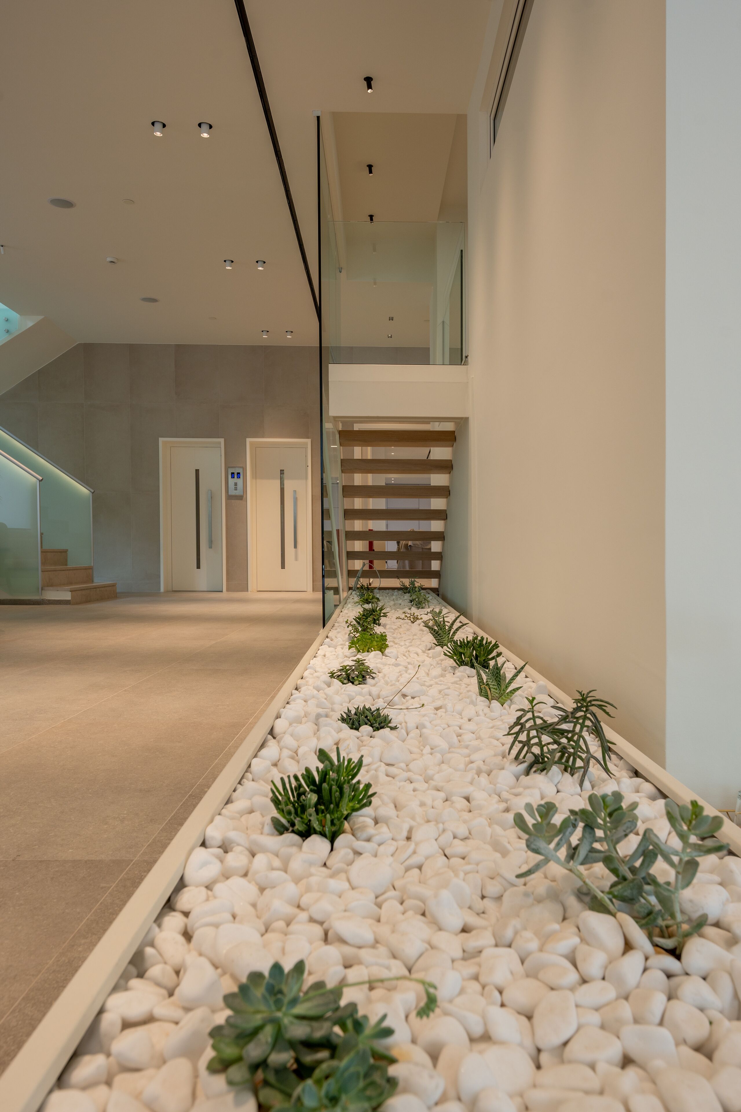 Un pasillo interior moderno con un sendero de guijarros, barandillas de vidrio y una escalera.
