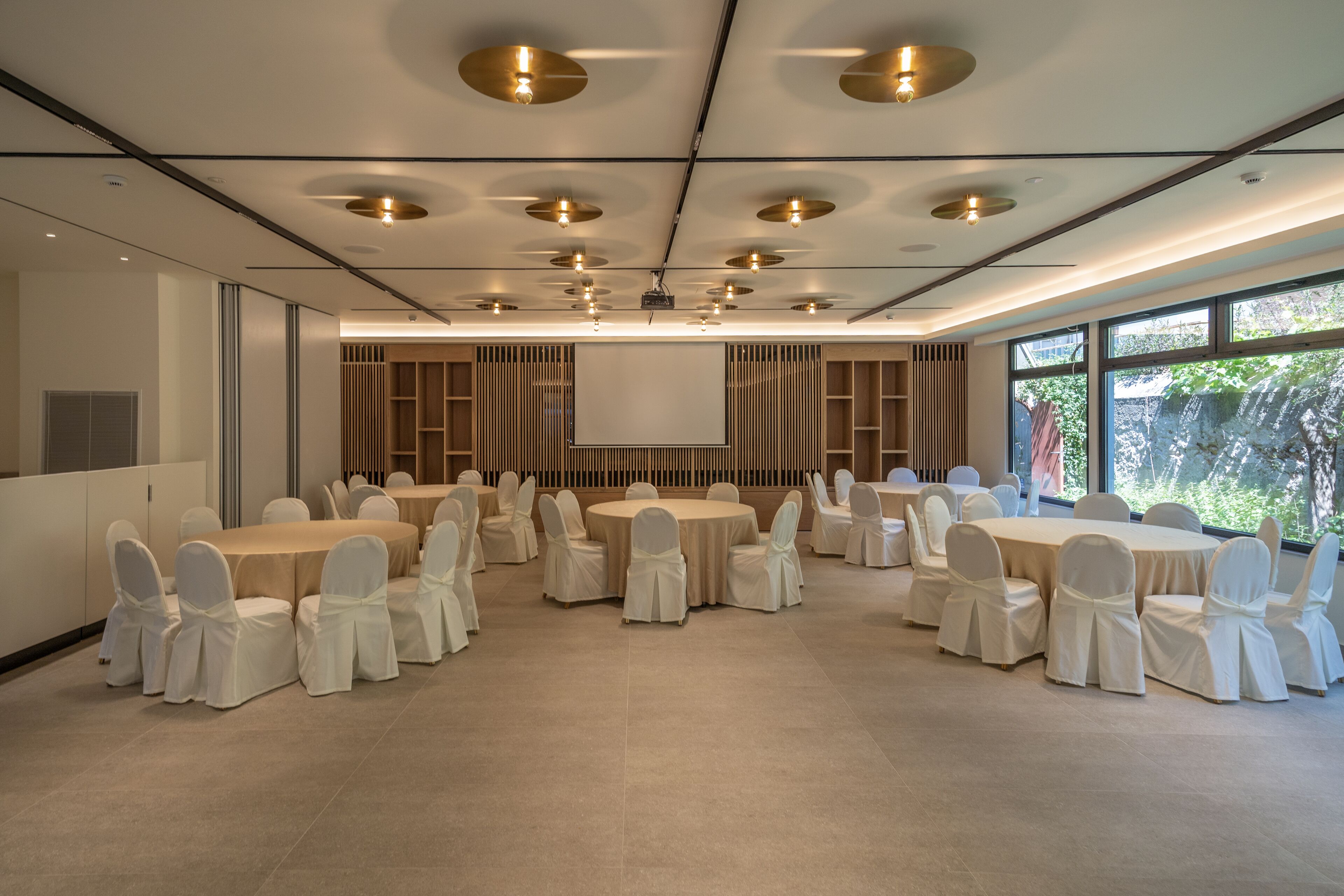 Una sala conferenze con tavoli e sedie rotonde, uno schermo per proiettore e ampie finestre.