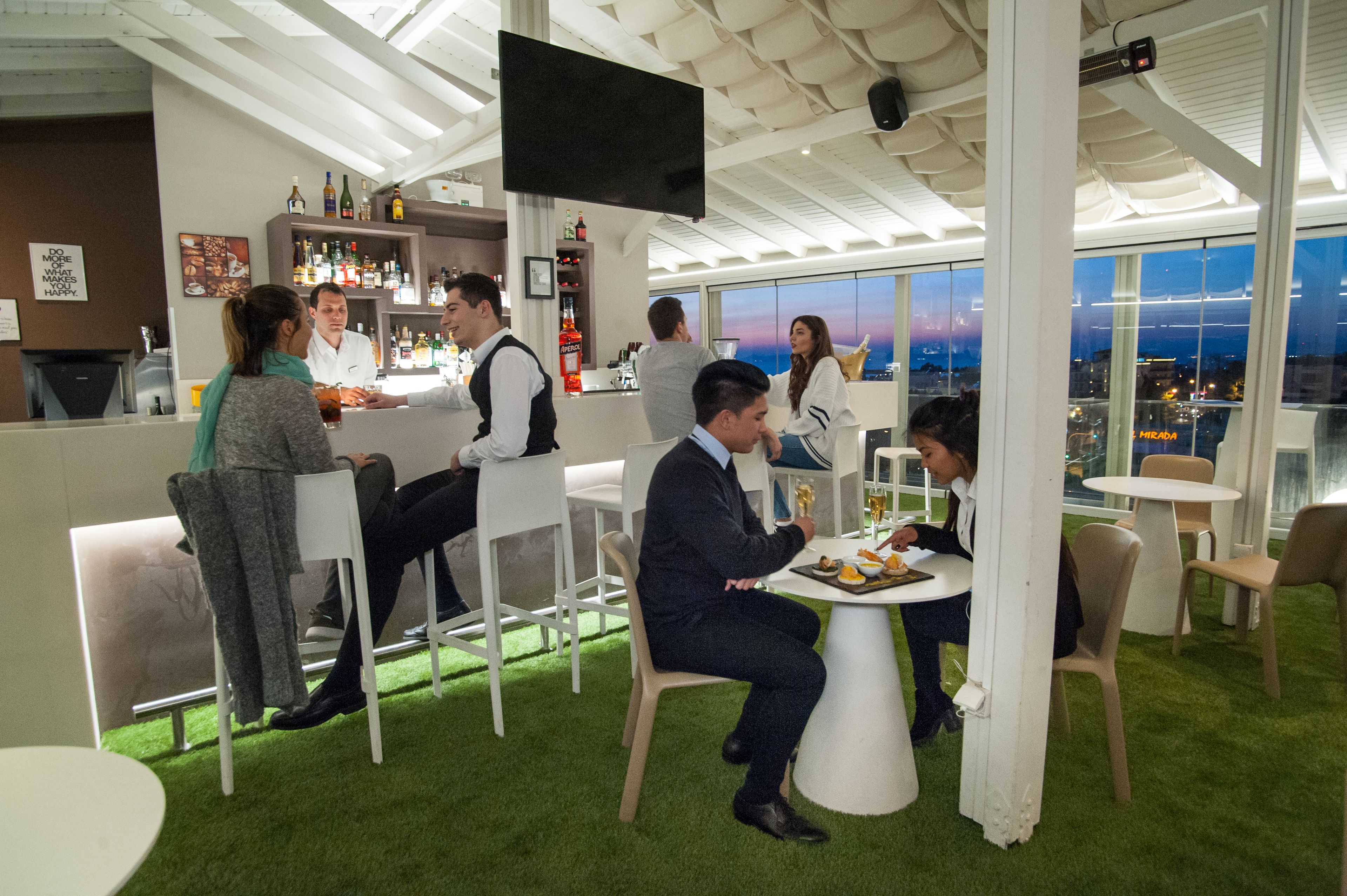 Eine moderne Bar mit weißen Möbeln, einer Bar und Blick auf die Stadt im Abenddämmerung.