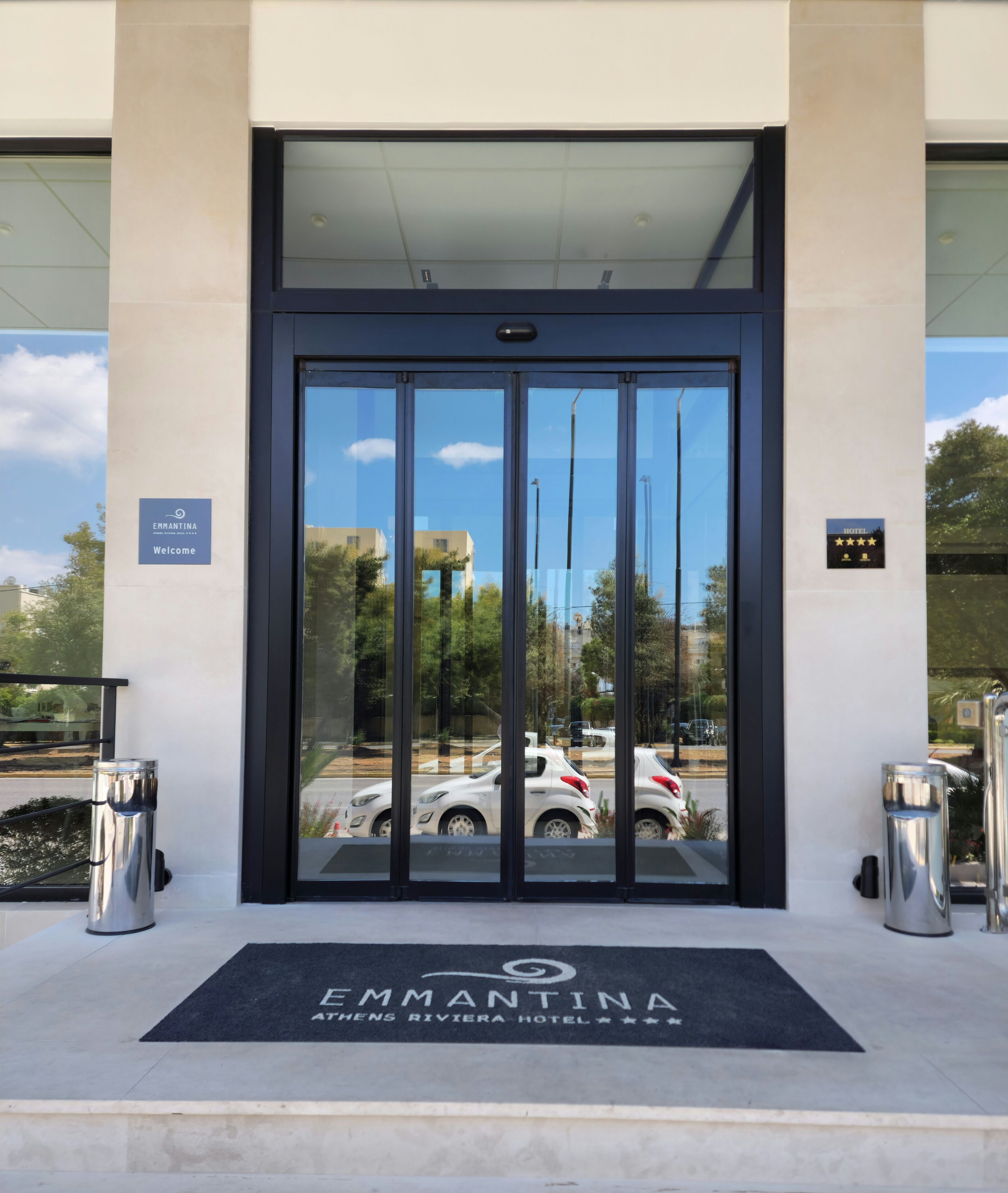 De ingang van het Emmantina Athens Riviera Hotel met een glazen deur, een welkomstbord en een deurmat.