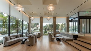Un área de estar moderna con sofá, sillas y mesas, grandes ventanales con vistas al exterior.
