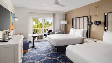 A hotel room with a bed, a desk, a chair, a table, a ceiling fan, and a view of palm trees.
