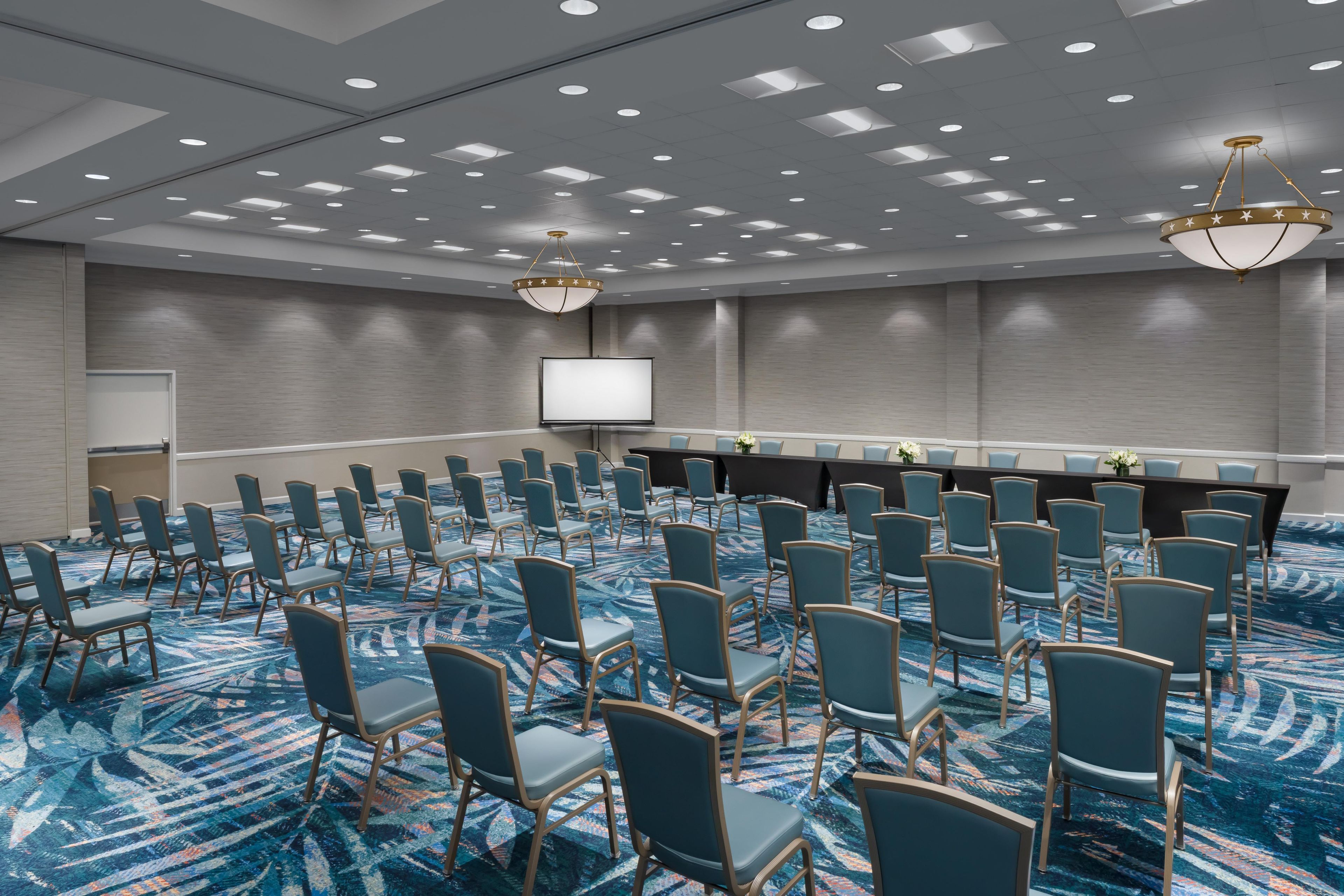 A conference room with rows of chairs, a large table, a projector screen, and a patterned carpet.