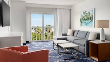 Une chambre d’hôtel moderne avec un balcon, un canapé gris, une table basse blanche, un fauteuil rouge et un tableau représentant une plage tropicale.