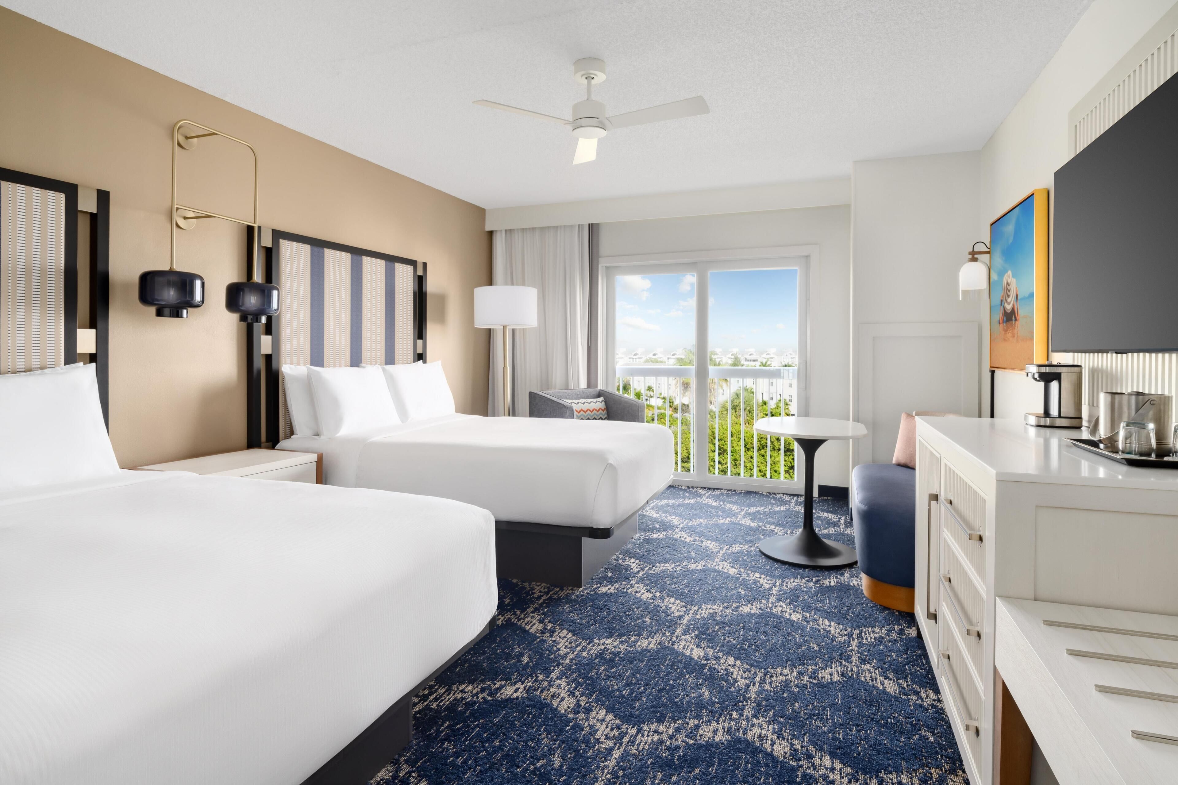 A hotel room with two beds, a desk, a chair, a small table, a ceiling fan, and a view of the outdoors.
