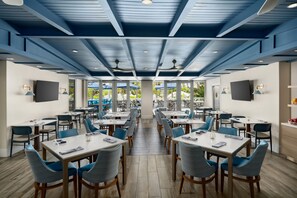 A modern dining area with multiple tables and chairs, large windows, and ceiling fans.