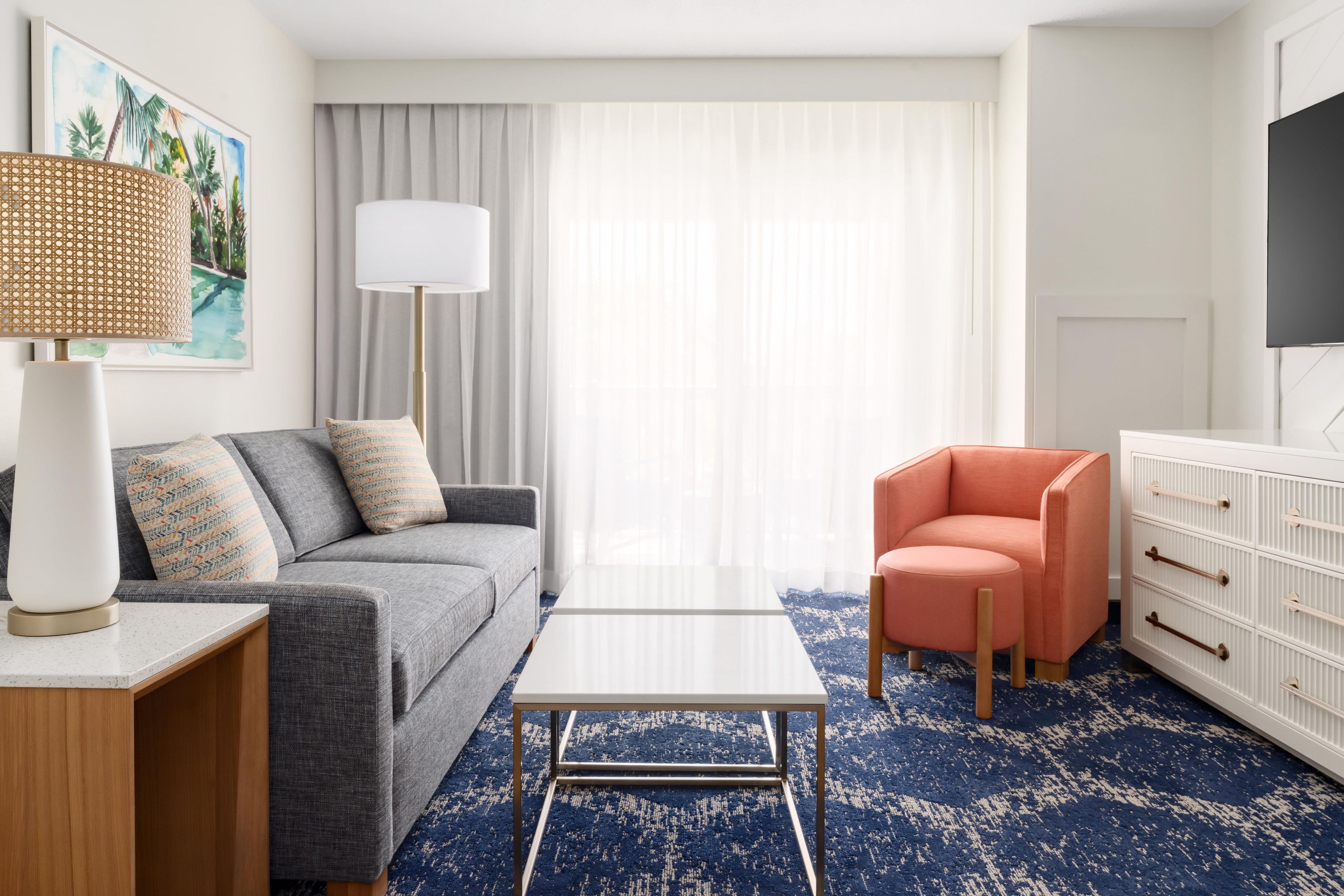 Un cuarto de hotel con un sofá gris, un sillón naranja pequeño, una mesita de centro blanca, un ventanal con cortinas translúcidas, una lámpara de pie y un televisor de pantalla plana montado en la pared.