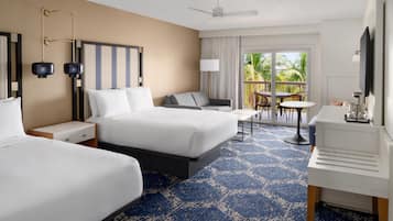 A hotel room with two beds, a desk, a chair, a TV, a ceiling fan, and a balcony with a view of palm trees.