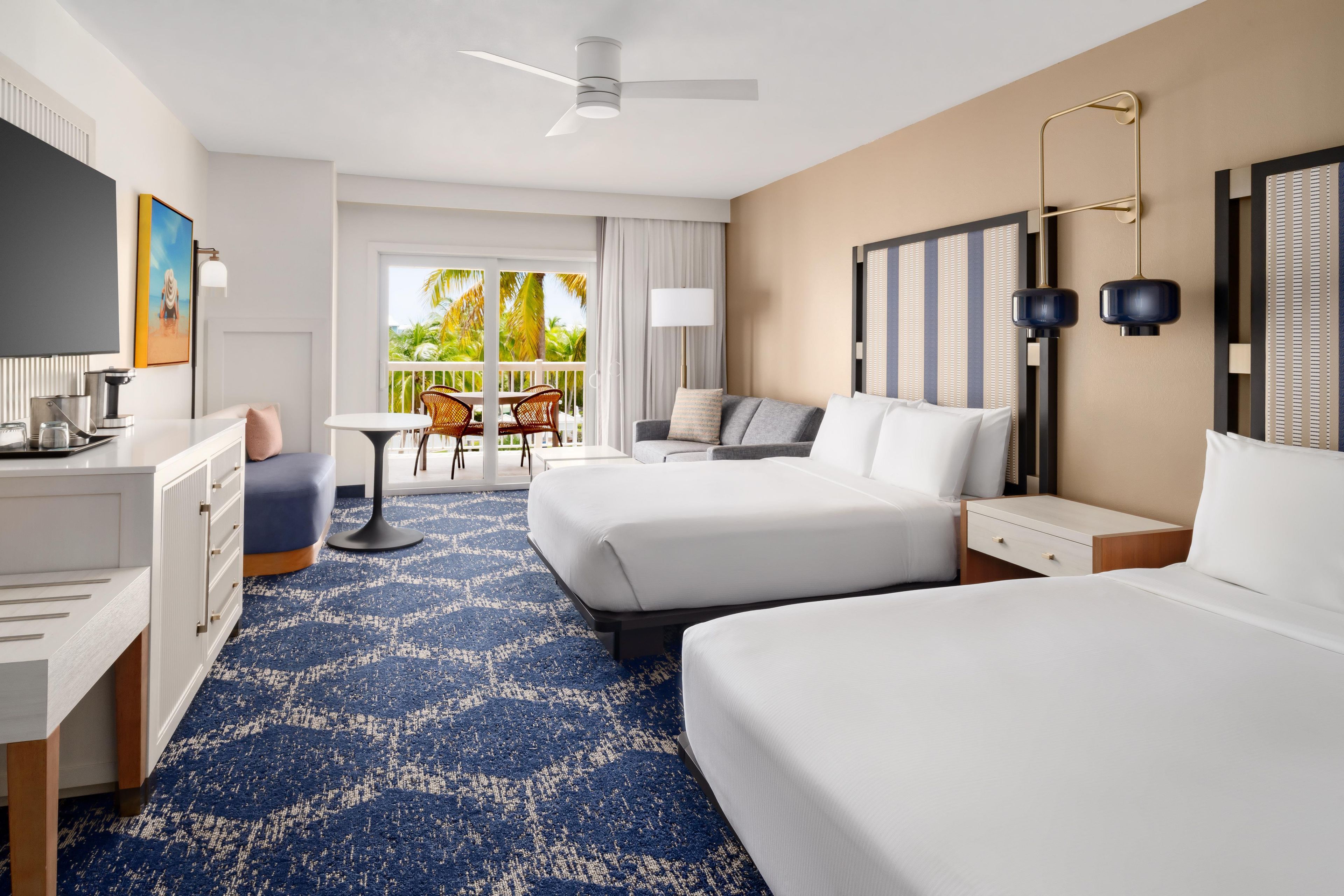 A hotel room with two beds, a desk, a chair, a balcony with a view, and a ceiling fan.