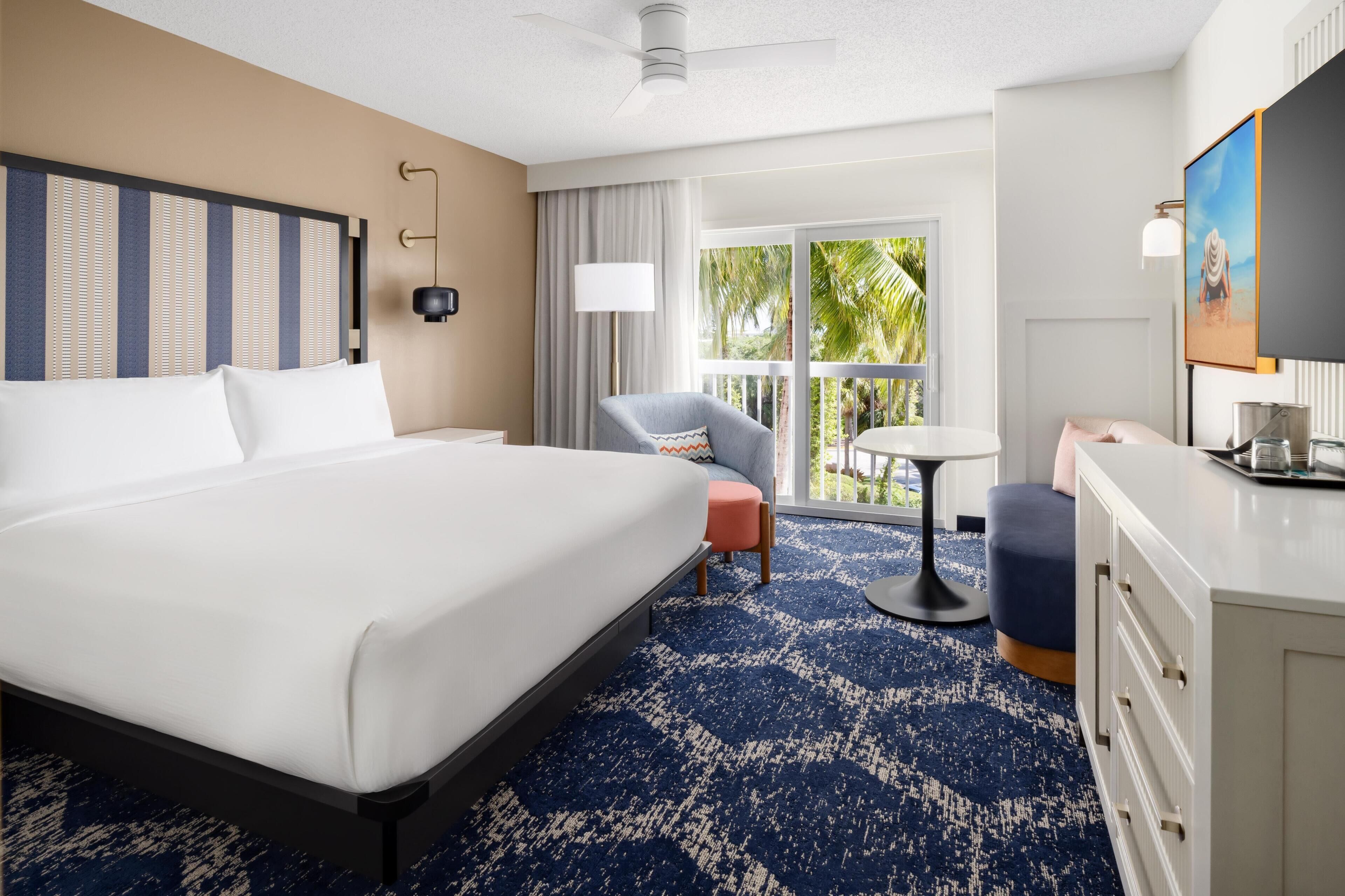 A hotel room with a large bed, a desk, a chair, a small table, and a balcony with a view of palm trees.