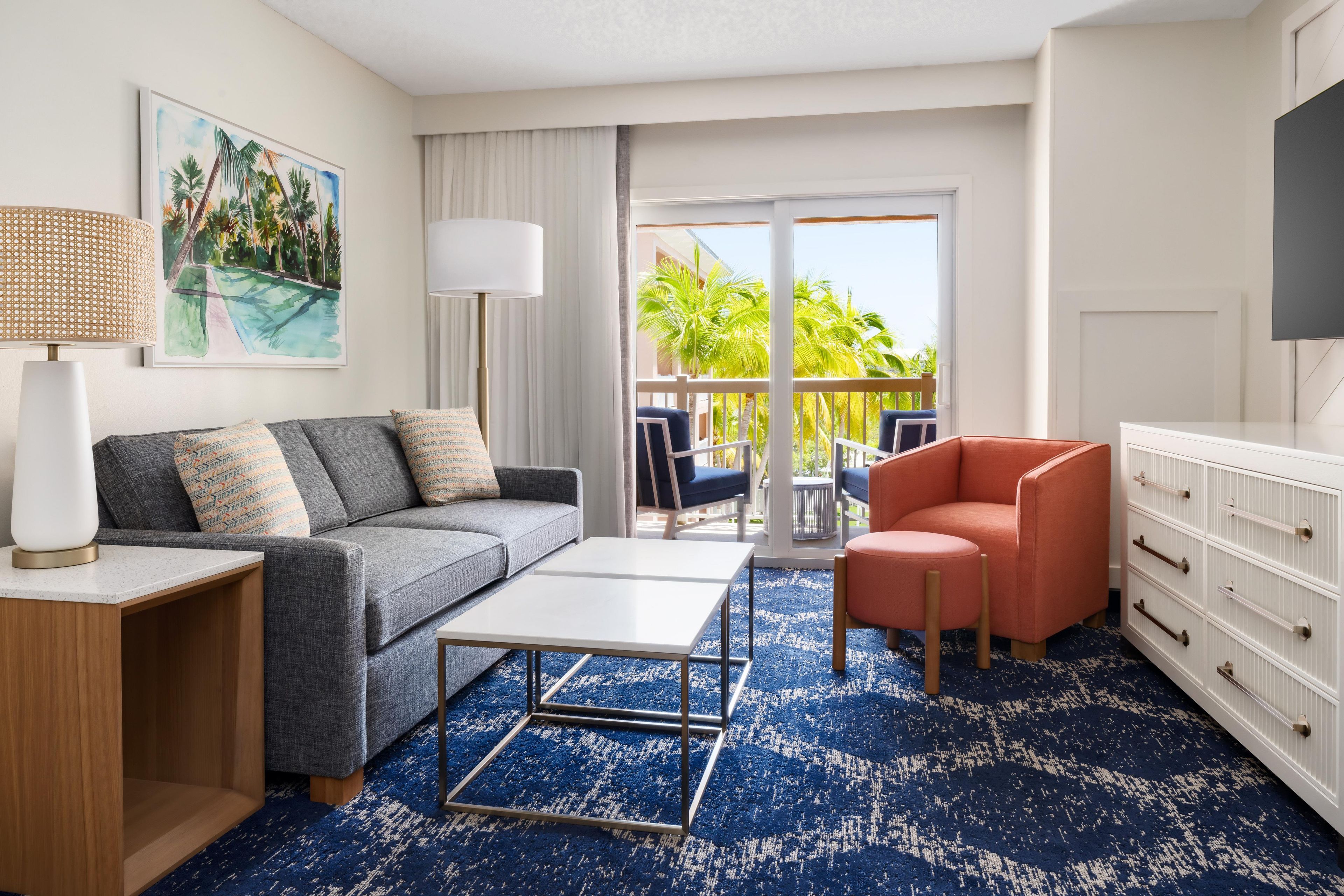 Una habitación de hotel moderna con un sofá gris, una mesa de centro blanca, un sillón rojo y un ventanal con vistas a palmeras.