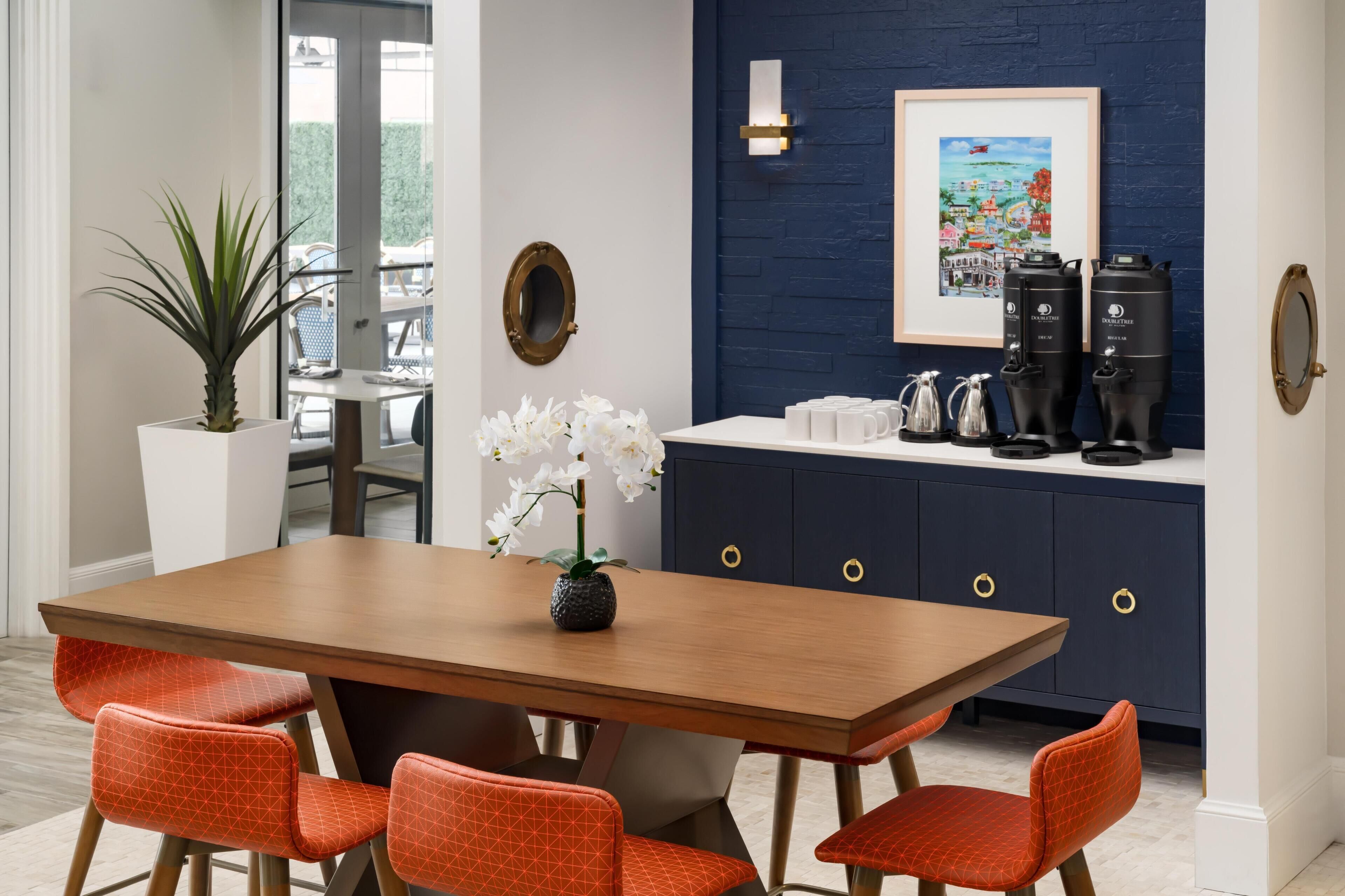 A dining area with a wooden table and orange chairs, a potted plant, white flowers, a framed picture, and a coffee station with two dispensers.