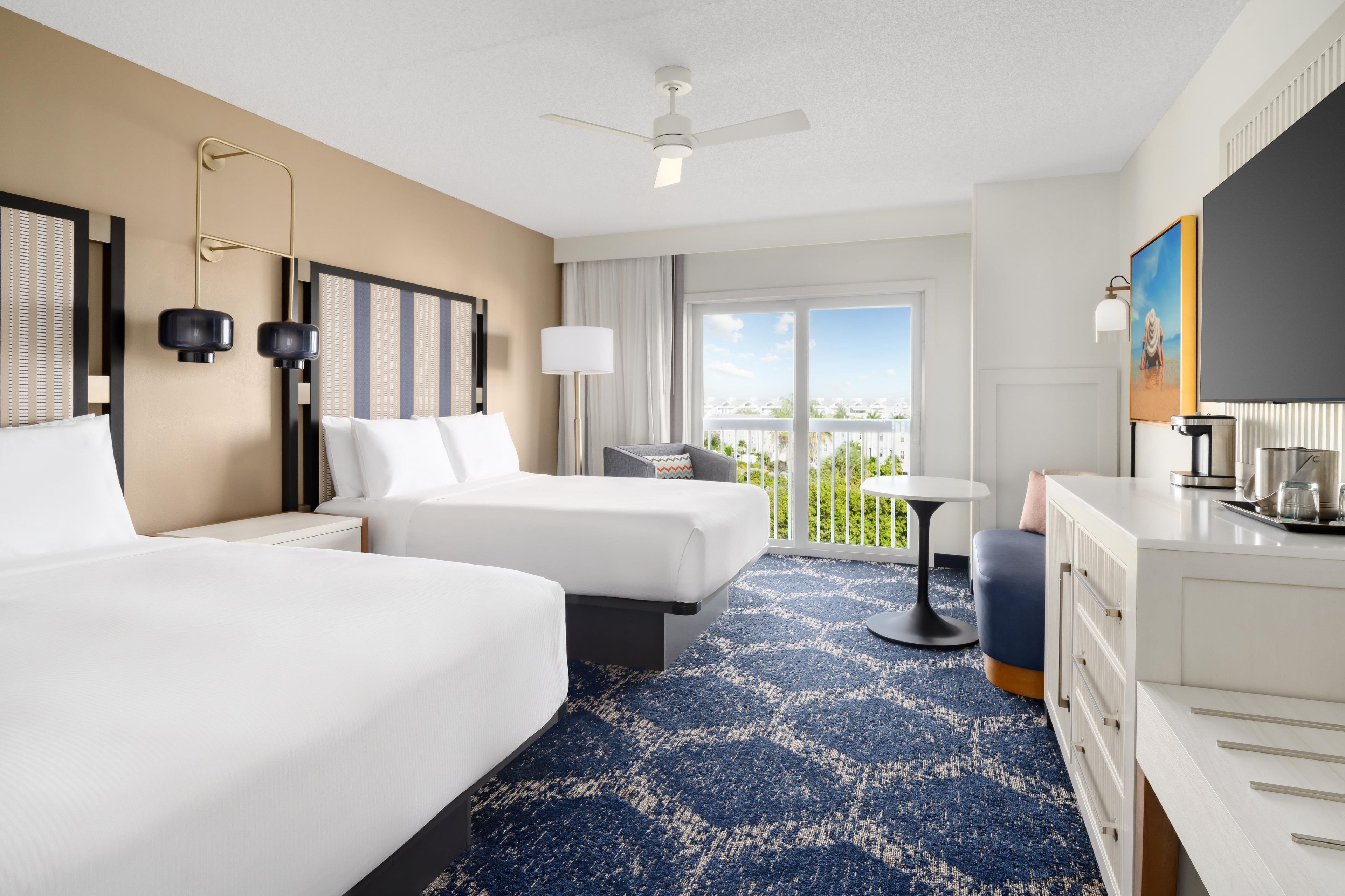 A hotel room with two beds, a desk, a chair, a balcony with a view, and a ceiling fan.
