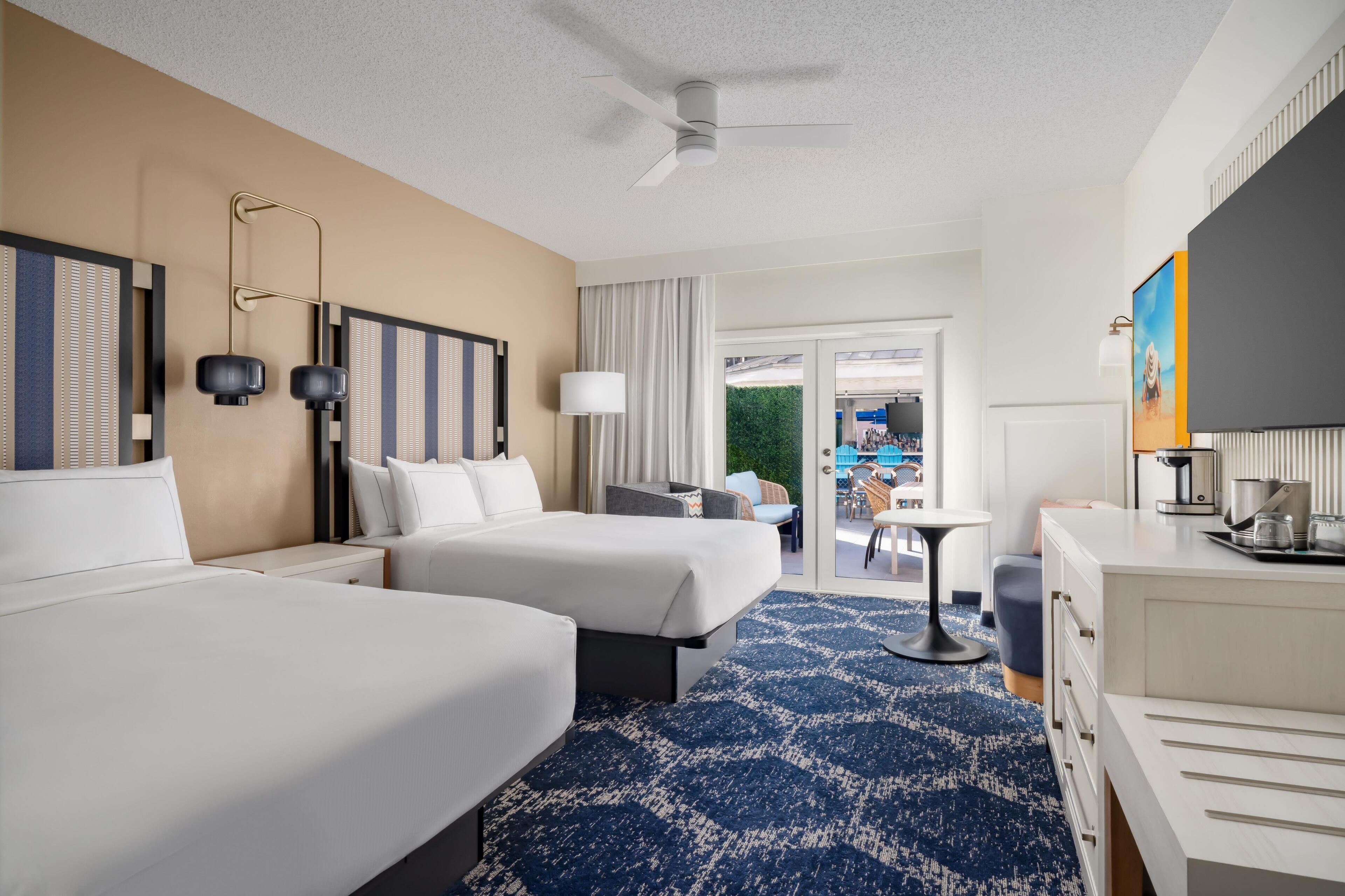 A hotel room with two beds, a desk, a chair, a coffee maker, and a view of the outdoor area.
