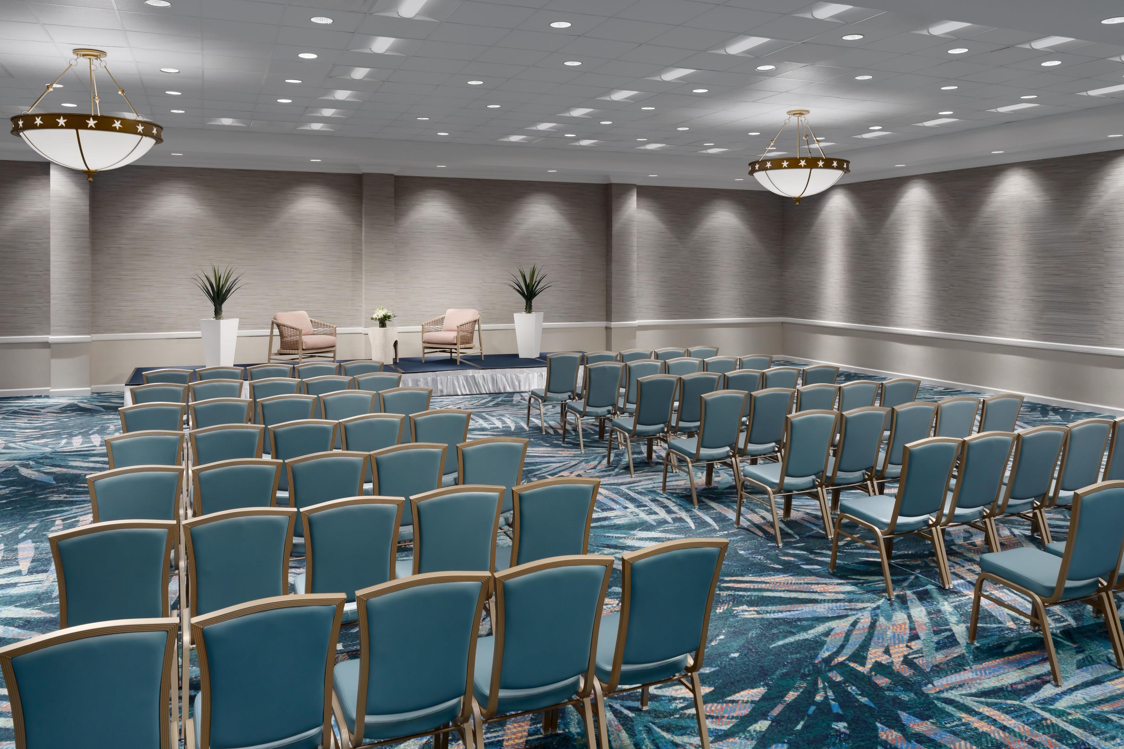 A conference room with rows of chairs, a stage with a table, and two chandeliers.