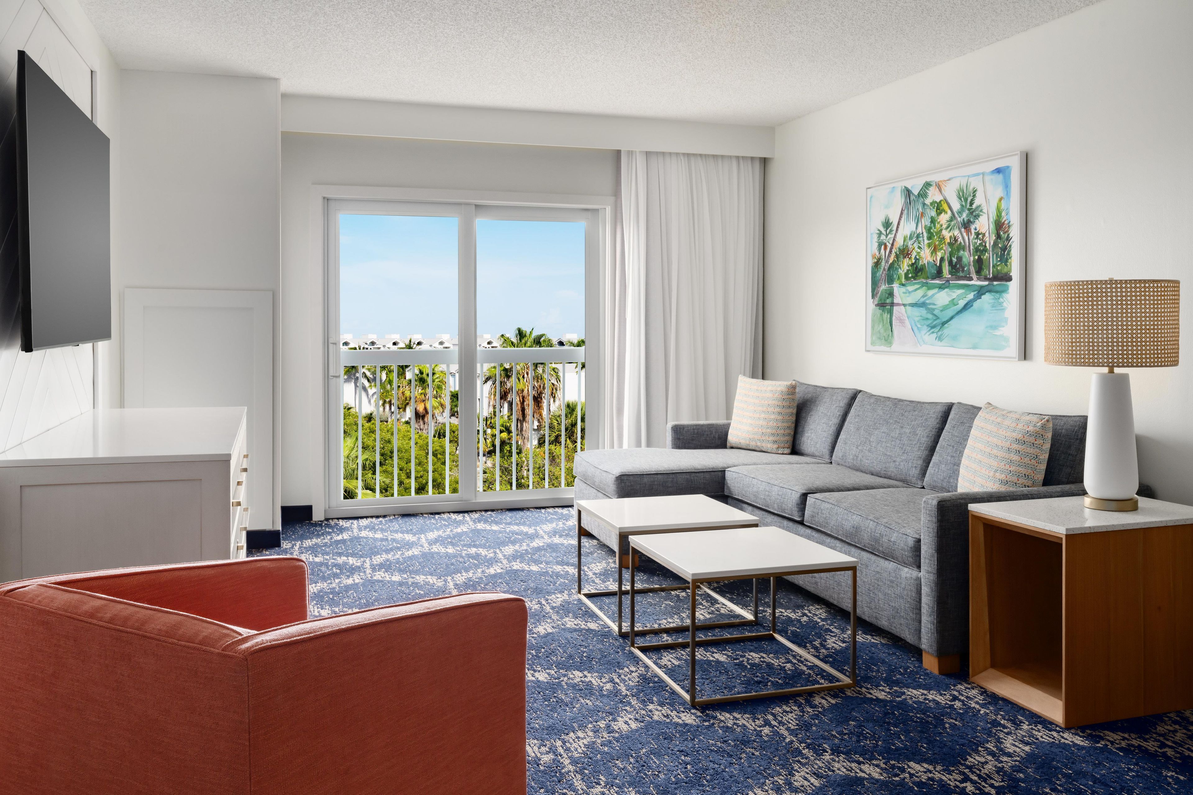 Quarto de hotel moderno com varanda, sofá cinza, cadeira vermelha, mesa de centro branca e pintura de uma paisagem tropical.